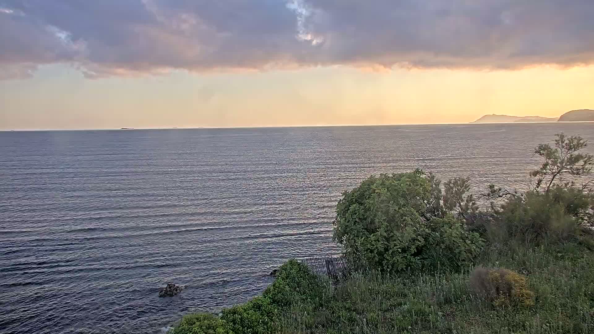 A calm sea stretches to the horizon under a partly cloudy sunset sky, viewed from a coastal vantage point with green vegetation in the foreground.
