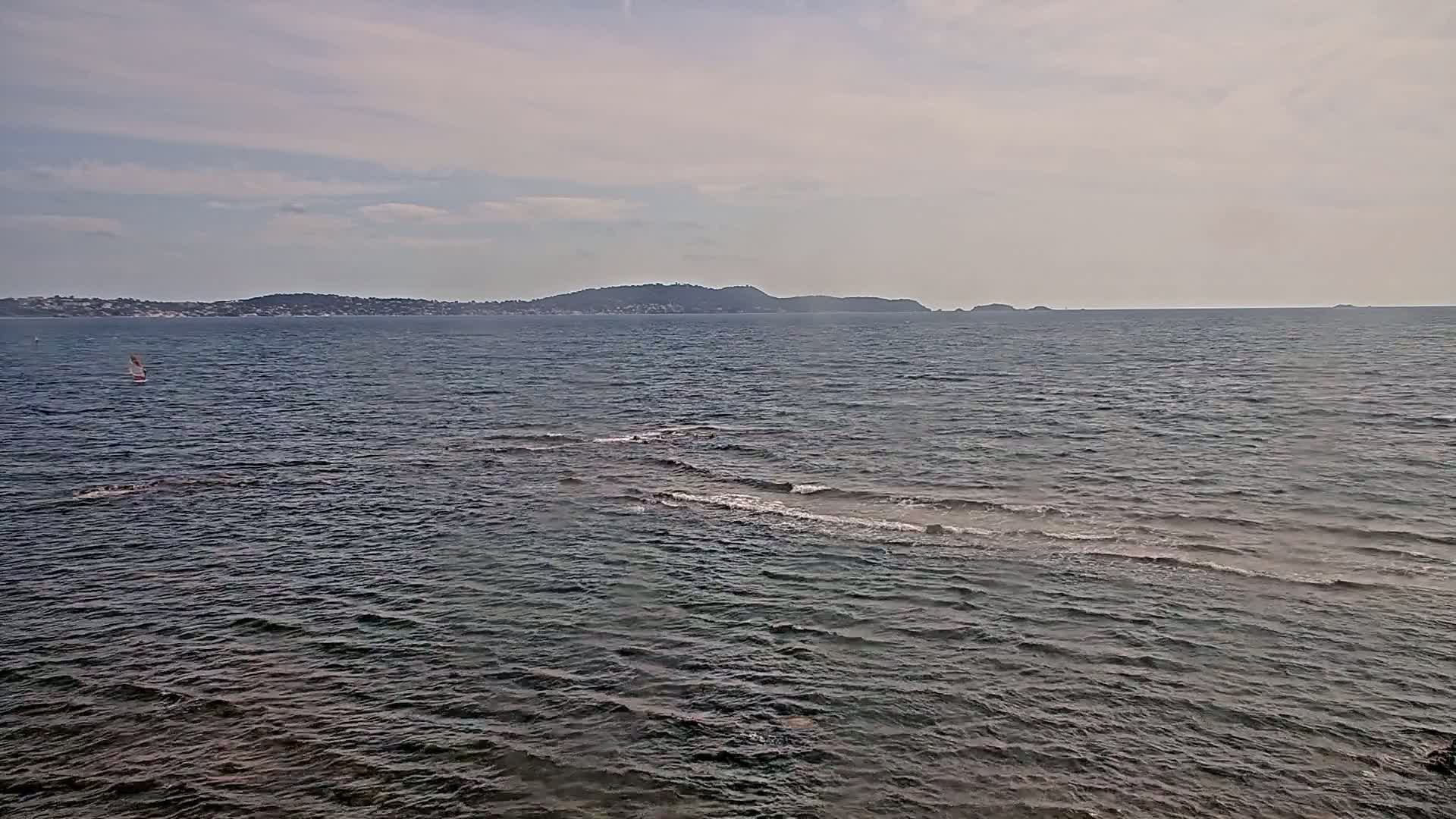A mostly calm sea with small waves shows a distant shoreline under a partly cloudy sky, and a single sailboat is visible on the water.