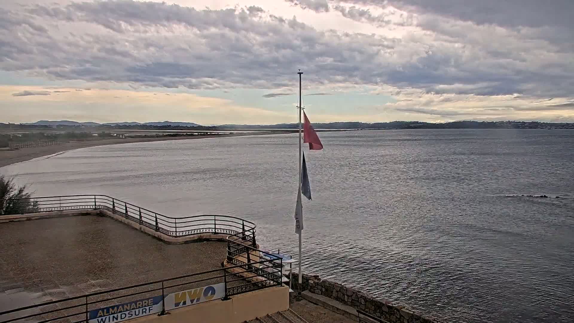 A mostly cloudy sky overlooks a calm body of water, a sandy beach, and a flagpole with several flags on a rooftop patio.