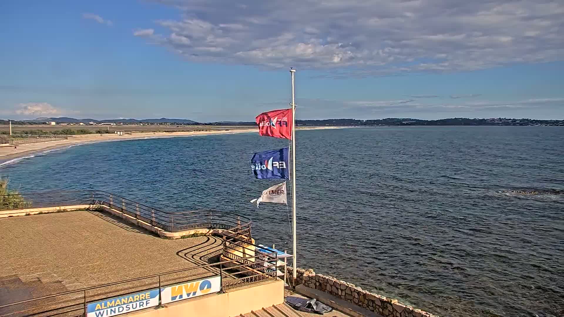 A partly cloudy day overlooks a sandy beach and calm ocean water, with three flags on a pole near a stone structure.