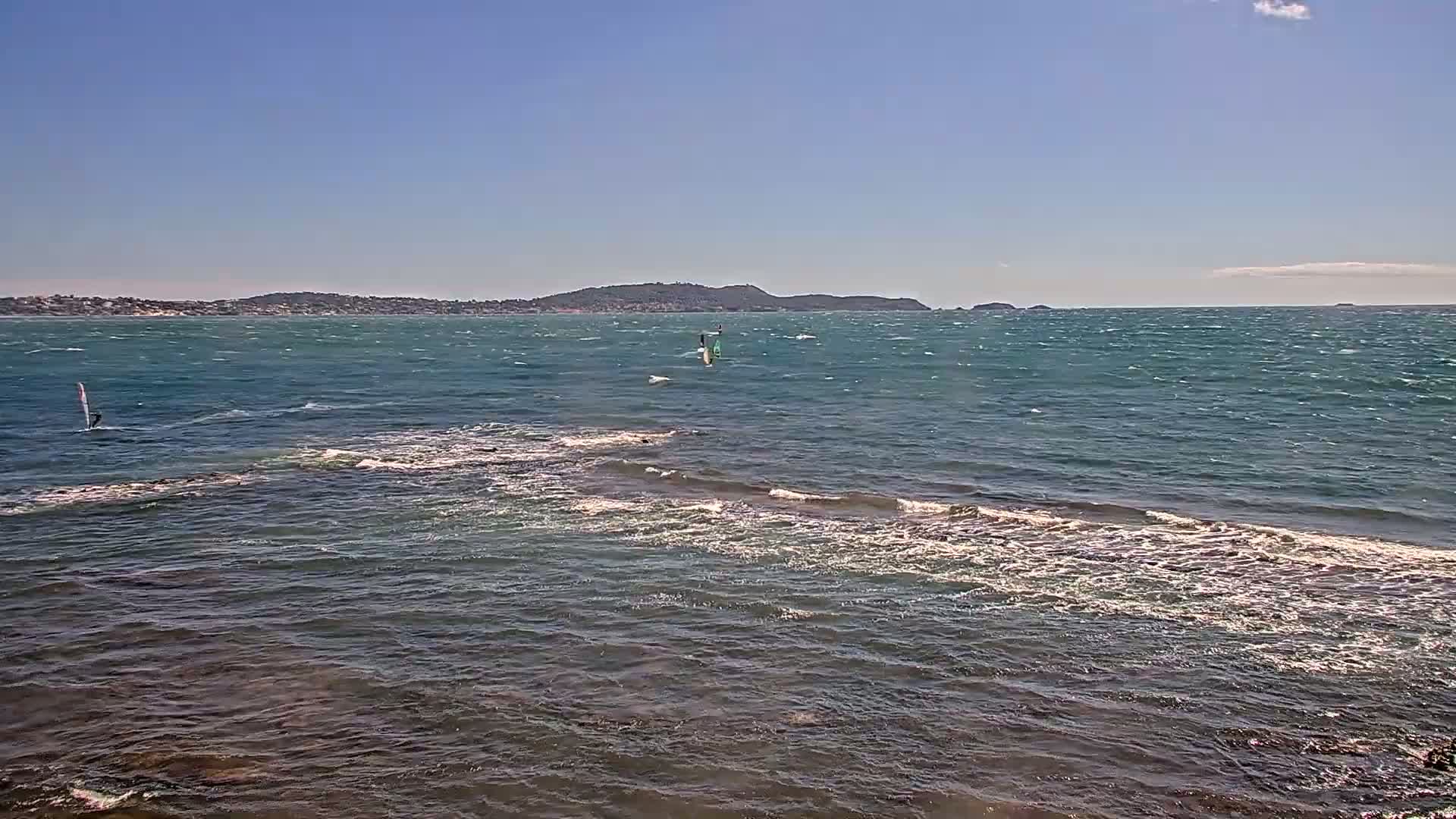 A partly sunny day shows wind-whipped water with two windsurfers visible near a rocky shoreline and a distant landmass under a clear sky.