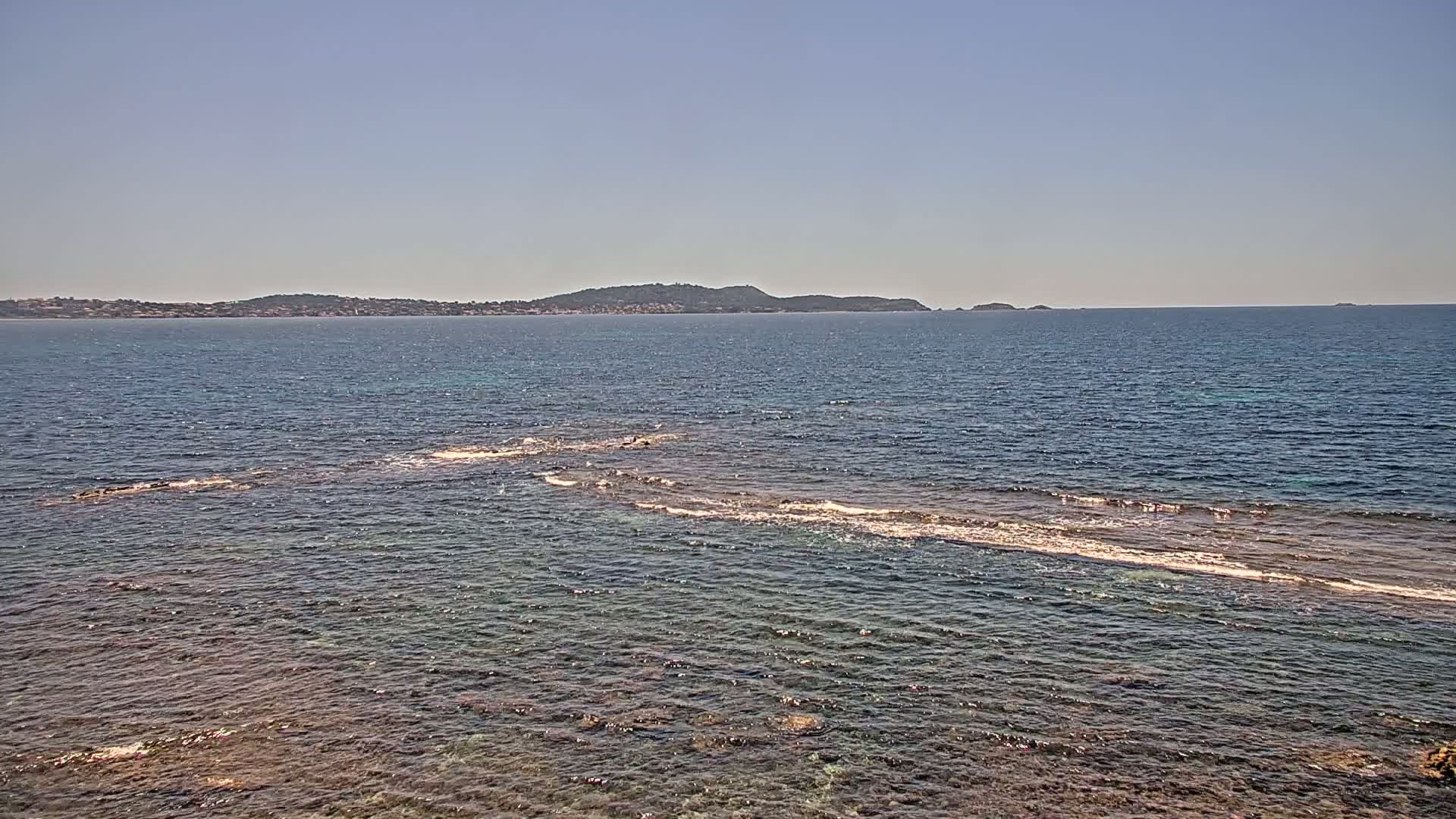 A calm, clear sea with a distant island and submerged rocks visible near the shore under a bright, sunny sky.