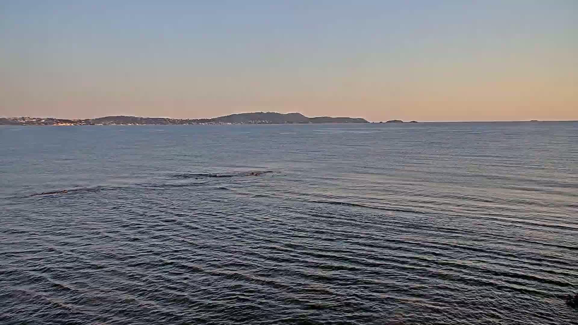 A calm sea with small waves under a clear, pale sky shows a distant shoreline with low hills.