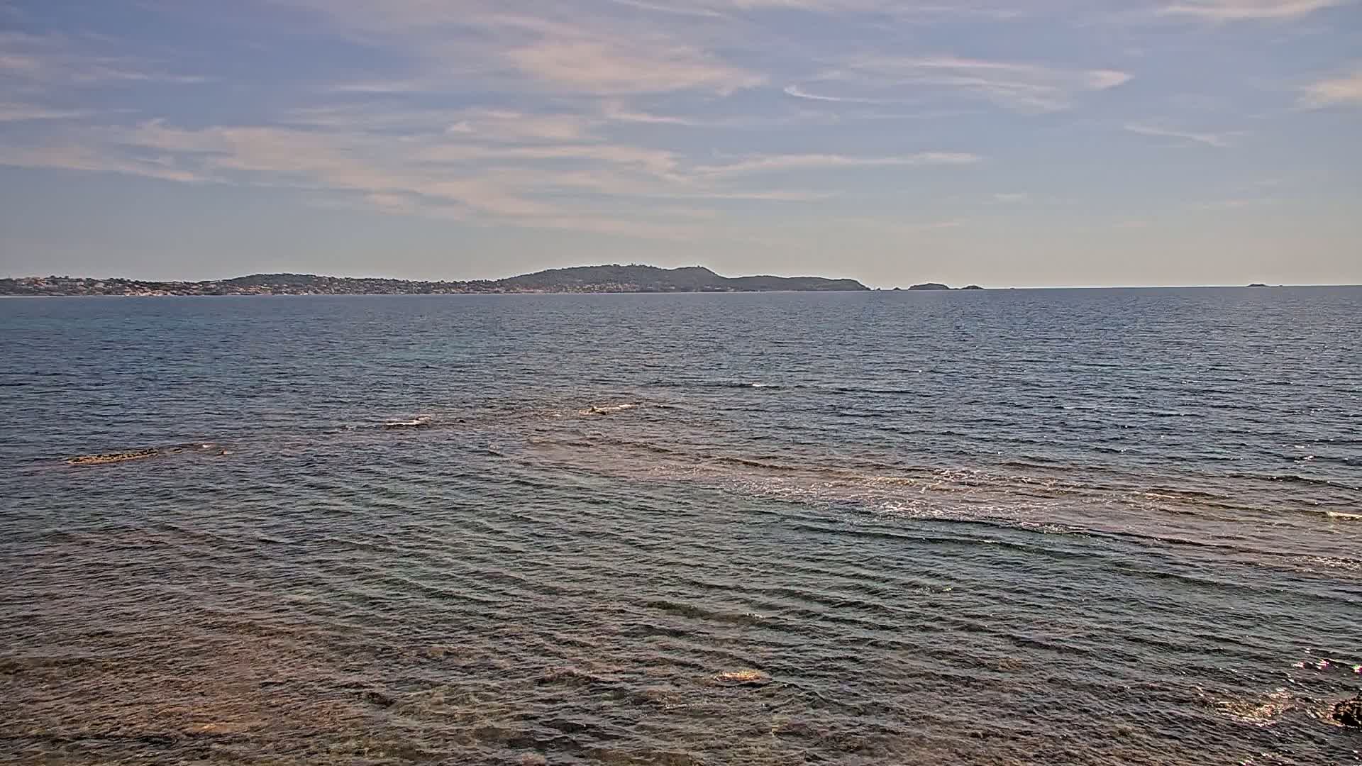 Calm, clear water laps gently against a rocky shoreline, with a distant landmass visible under a mostly sunny sky.