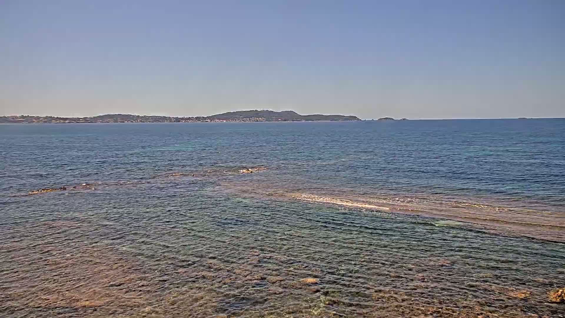 A calm, clear ocean with a rocky shoreline extends to a distant coastline under a clear, sunny sky.