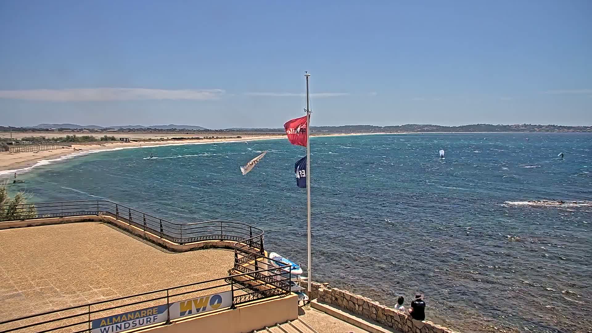 A sandy beach and wavy ocean are viewed from an elevated vantage point on a sunny day, with several windsurfers on the water and two people sitting on a low wall.