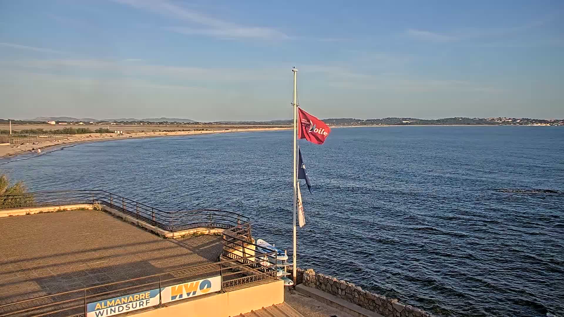 A sandy beach stretches along a calm ocean under a clear sky, with a flagpole bearing a red flag and other flags situated on a paved area overlooking the water.