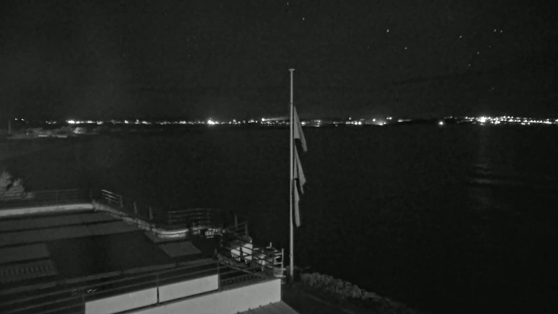 A nighttime view of a calm body of water with a distant shoreline of lights, a flagpole with furled flags, and a rooftop structure in the foreground.