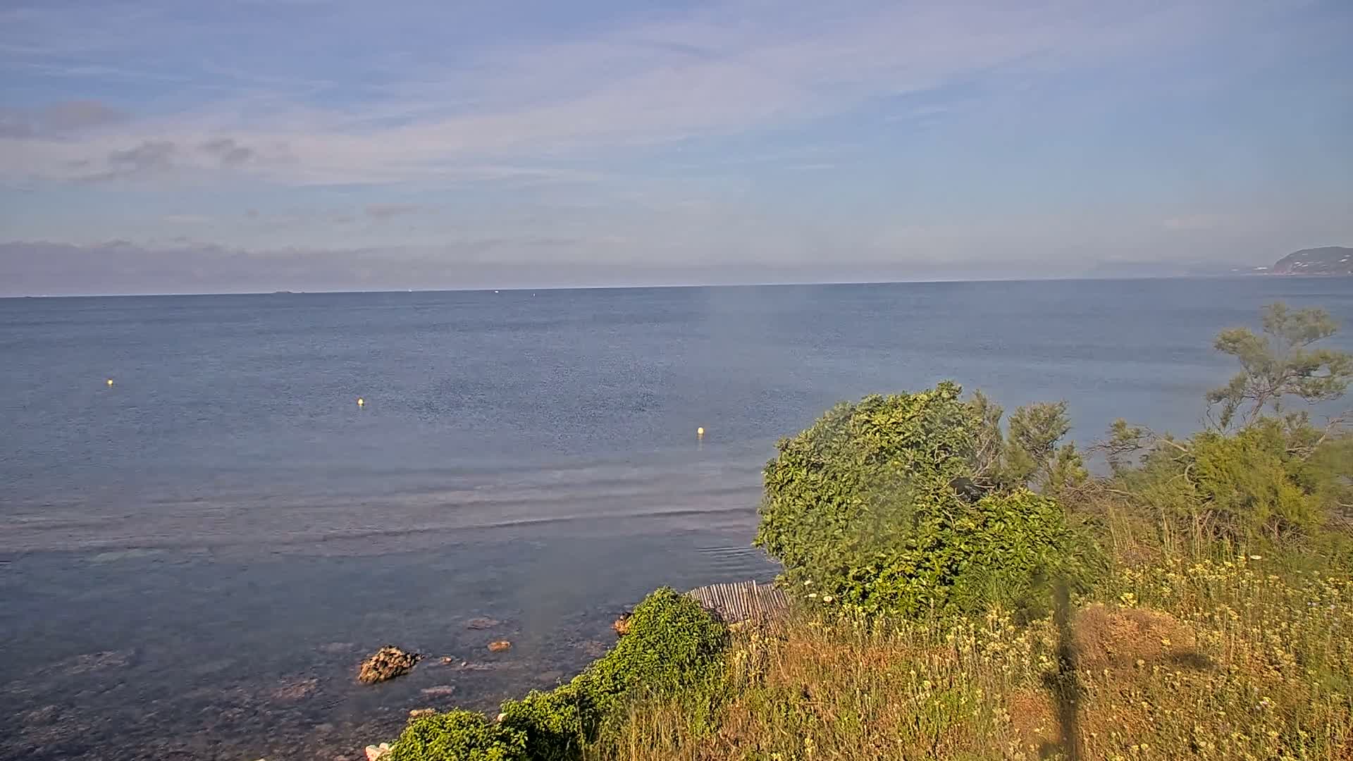 A calm, blue sea stretches to a distant shoreline under a mostly clear sky, viewed from a high vantage point overlooking a rocky coast with low-lying vegetation.