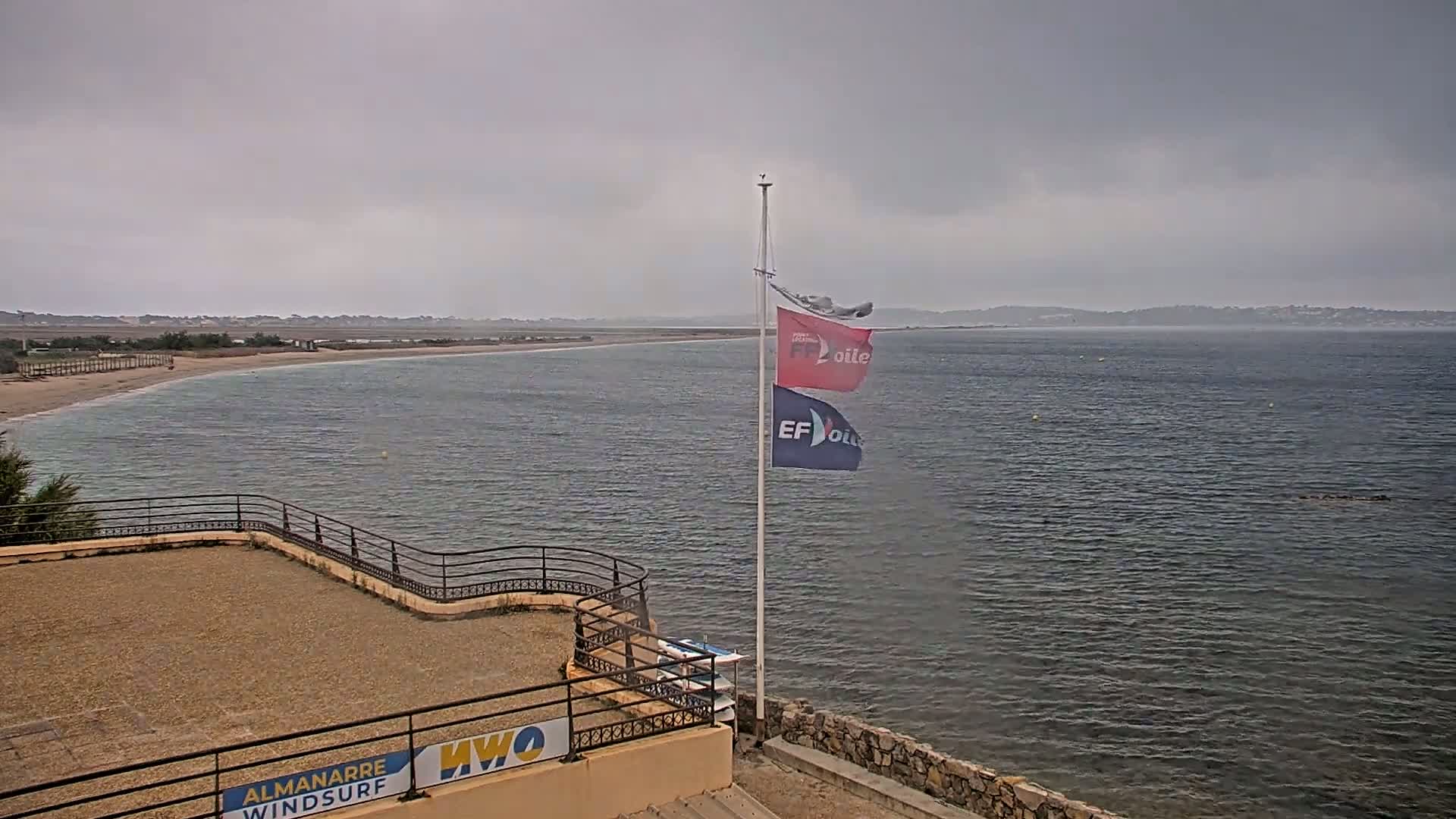 A sandy beach curves into a calm, gray sea under an overcast sky, with two flags on a pole in the foreground near a stone wall and small building.