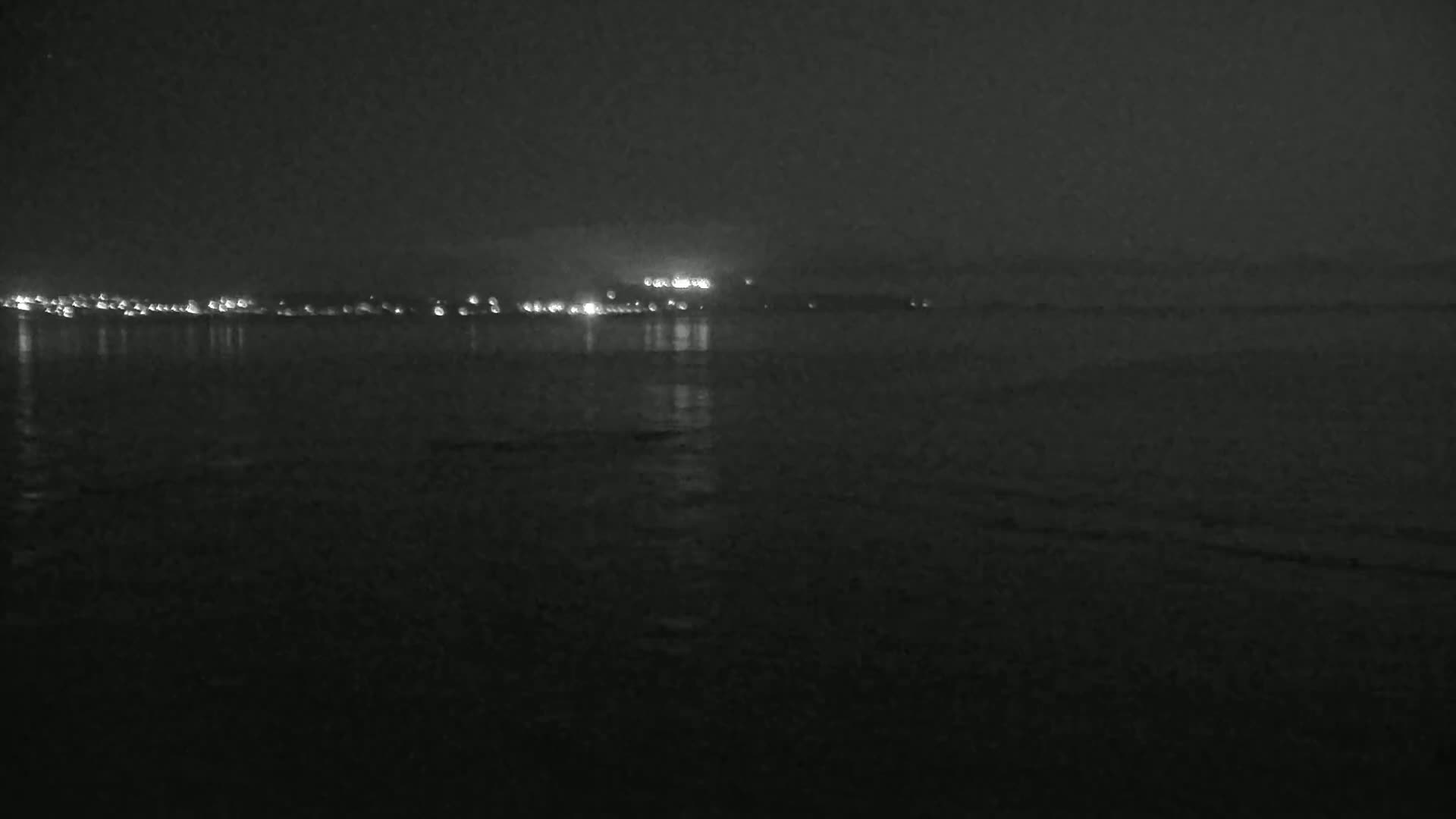 A dark, nighttime view of a calm body of water reflecting the lights of a distant city under a hazy sky.