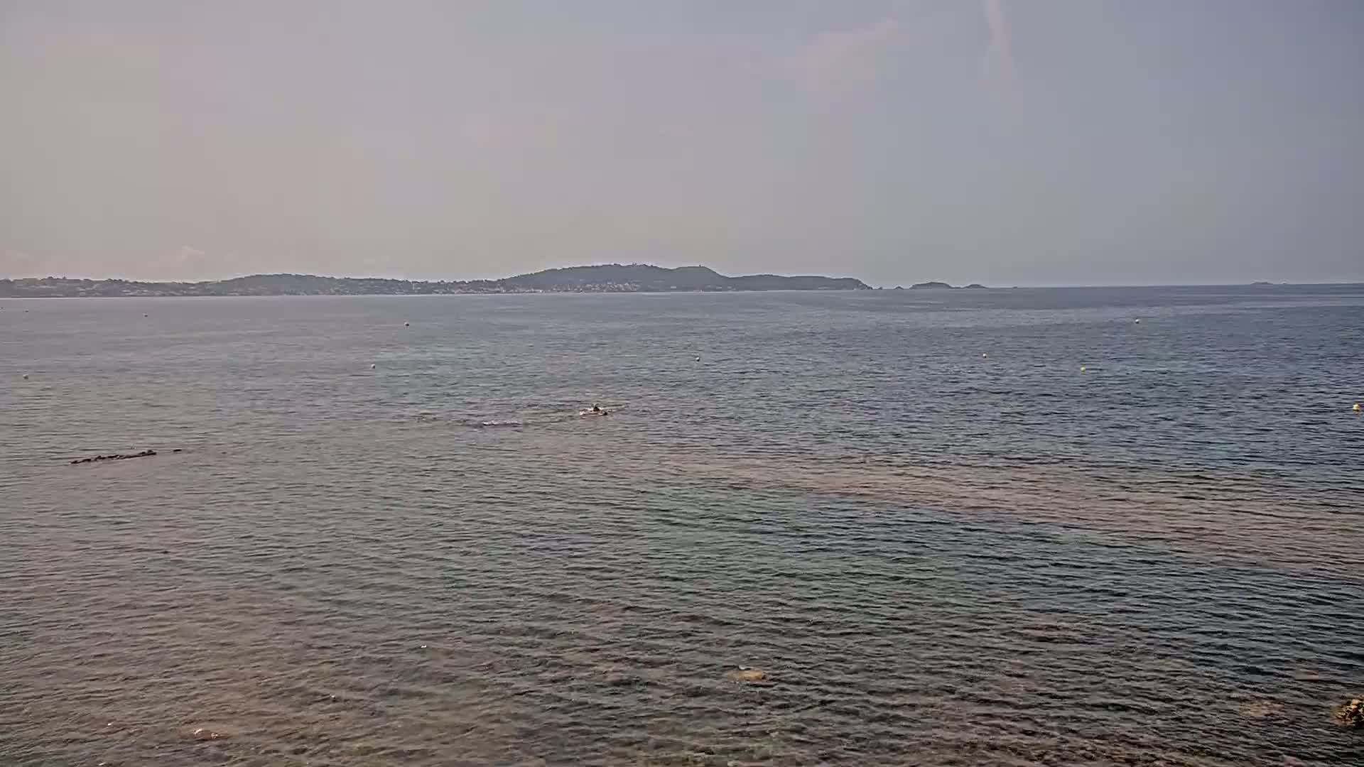 A calm, hazy sea stretches to a distant shoreline of low hills under a pale sky.