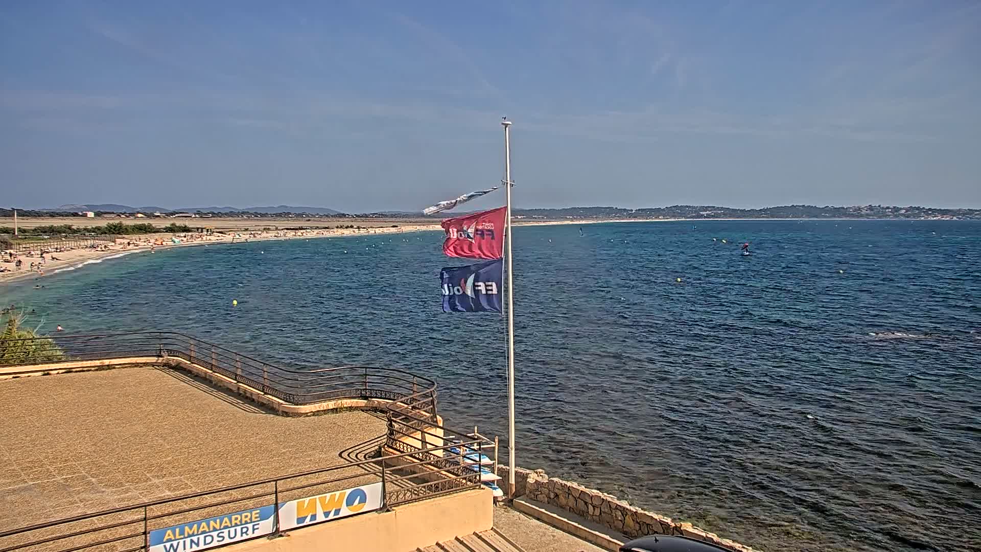 A sandy beach with people on it is next to a calm, blue ocean; three flags are on a pole near a stone structure overlooking the water on a sunny day.
