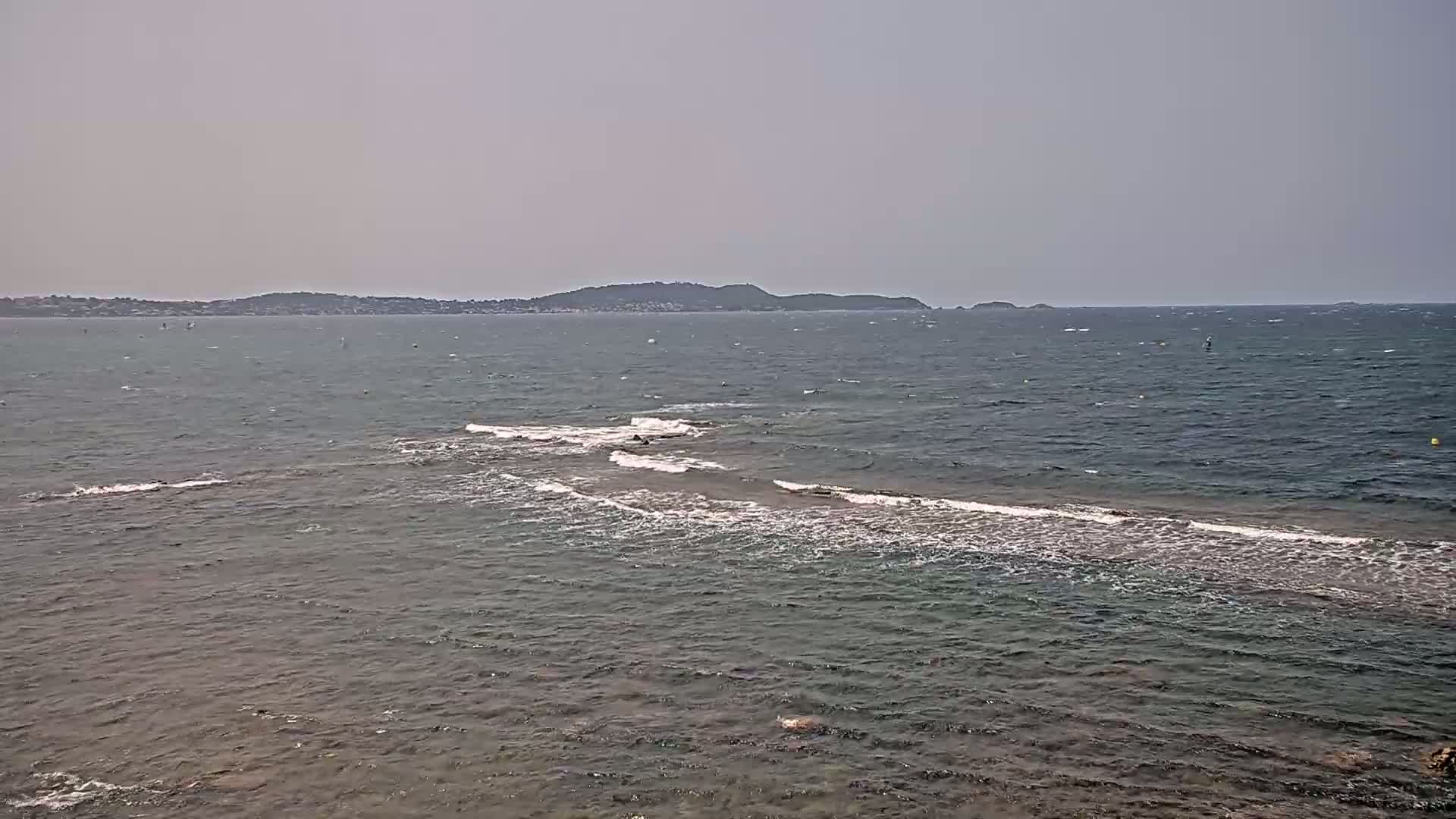 A partly sunny day shows a rocky shoreline with small waves lapping against it, a distant landmass visible across a choppy sea.