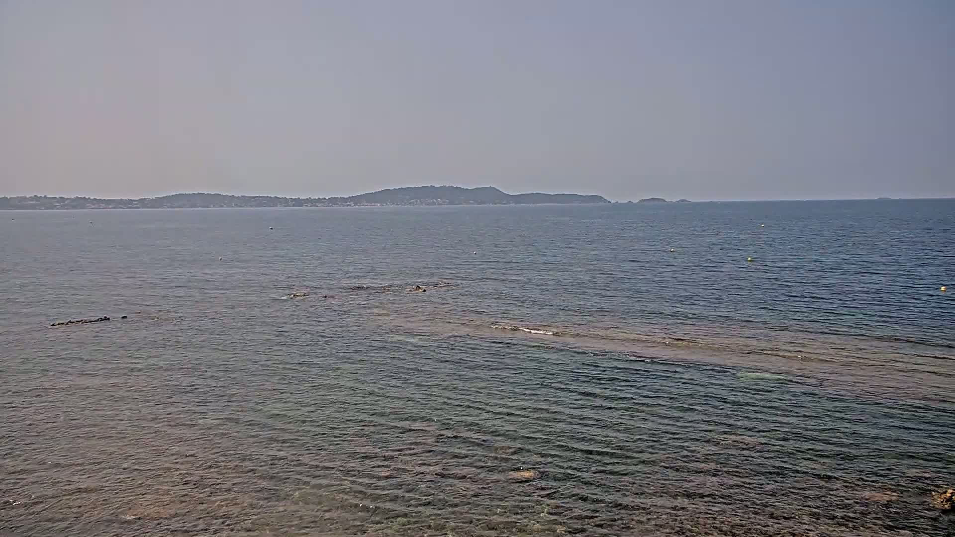 A calm, hazy sea with a distant mountain range under a light-grey sky shows shallows near the shore with dark rocks partially submerged.