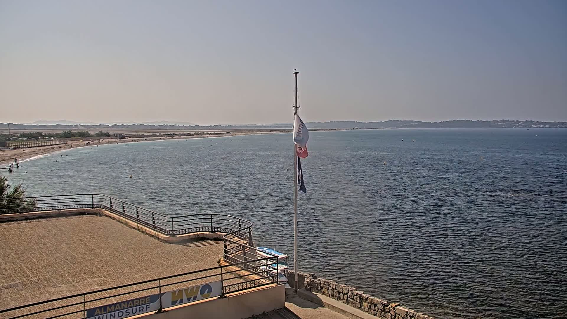 A sandy beach with a few people on it is visible next to calm water, with a distant shoreline under a clear sky.