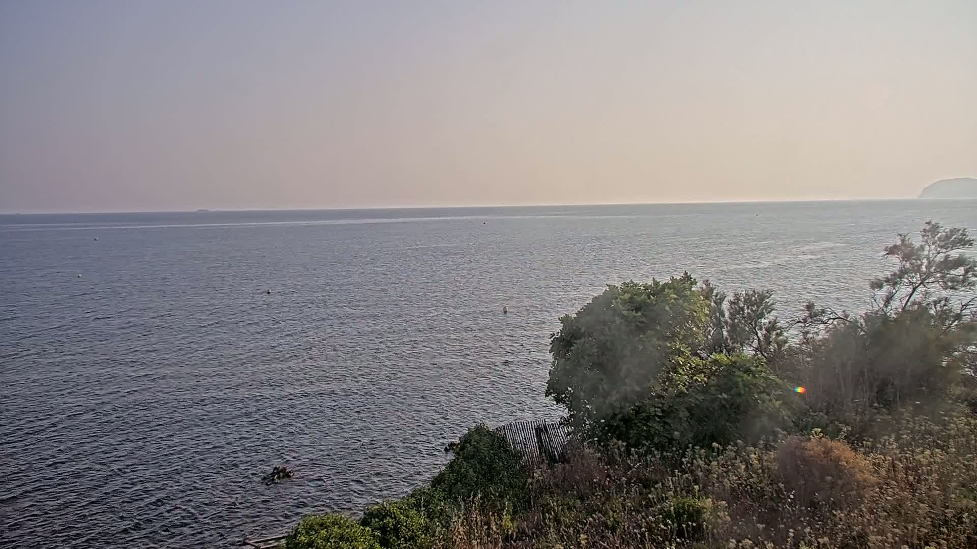 A calm, hazy sea is viewed from a high vantage point overlooking a shoreline with vegetation.