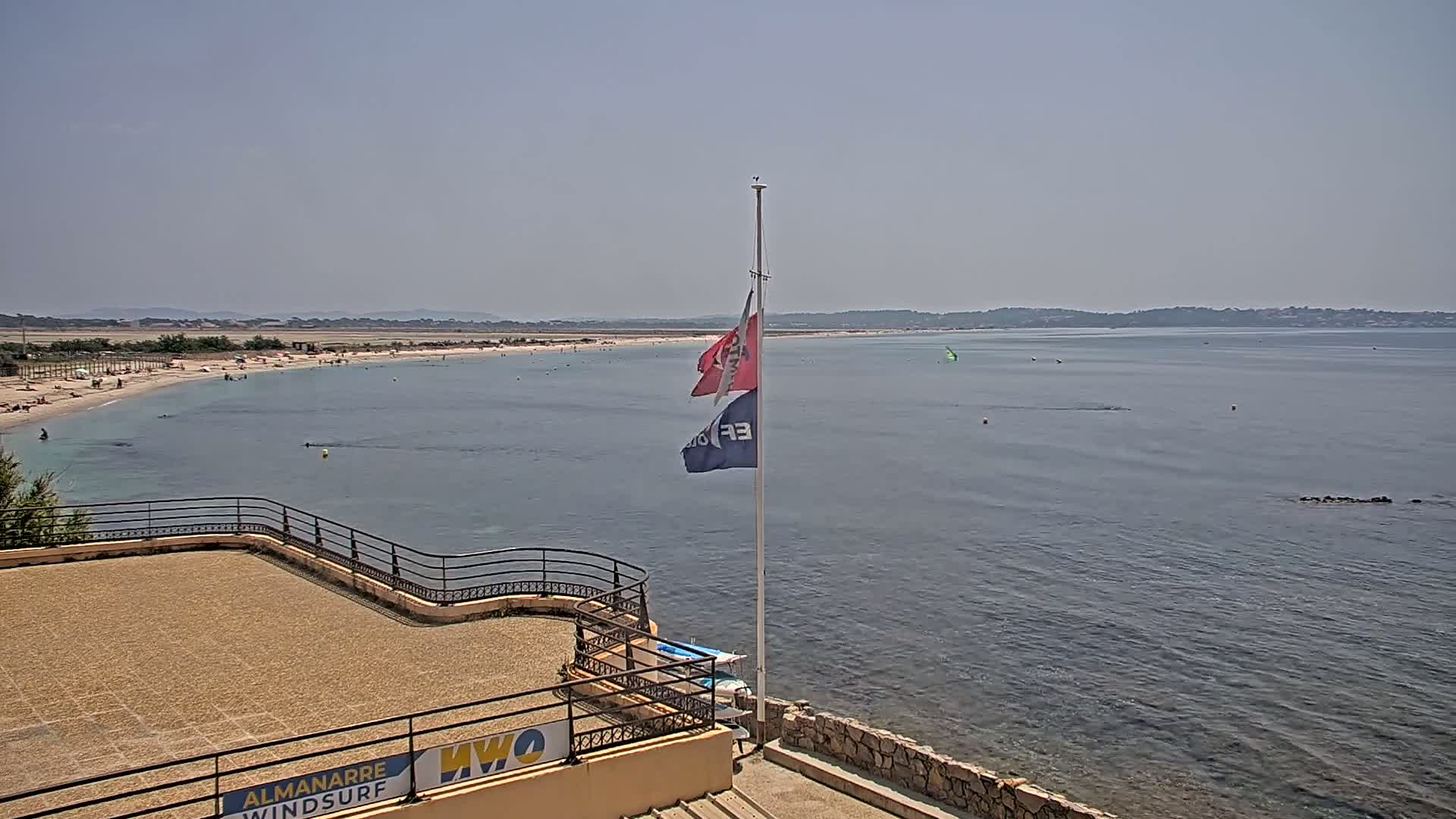 A sandy beach with scattered people is visible across a calm body of water, with two flags on a pole in the foreground under a clear sky.