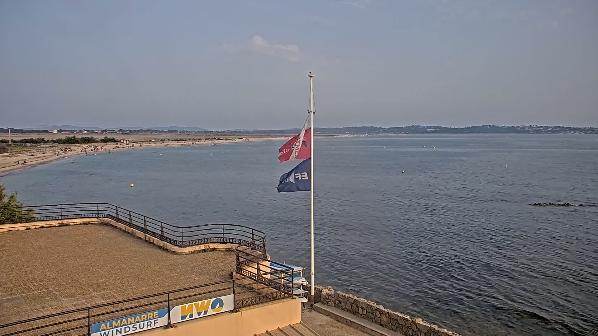 A sandy beach with scattered people stretches along a calm, blue body of water under a clear sky, and two flags fly from a pole near a low stone wall.