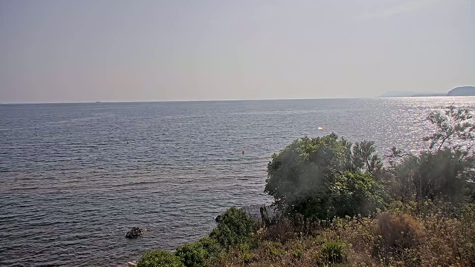 A calm, sunny day is shown overlooking a dark blue ocean with a distant shoreline and vegetation in the foreground.