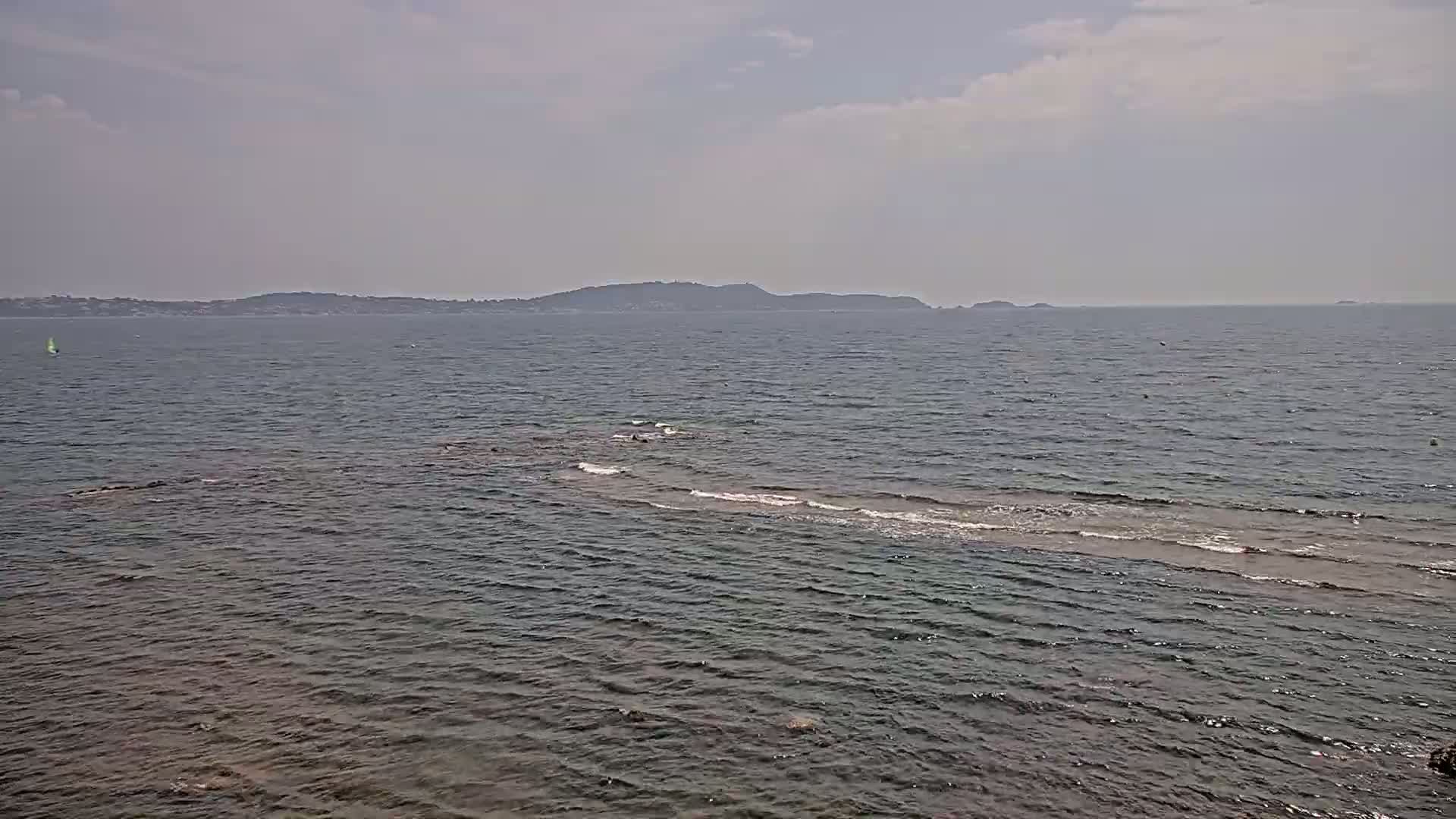 A calm sea with a distant shoreline under a partly cloudy sky shows a small, light-colored sandbar extending from the shore.