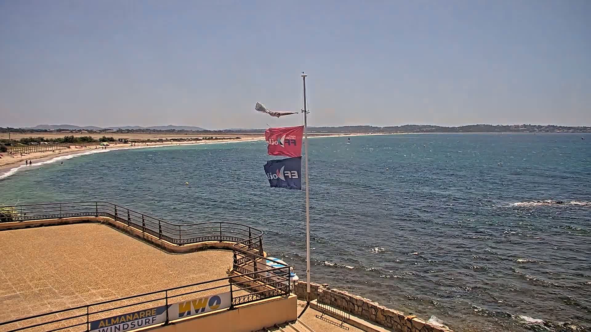 A sandy beach with a few people on it stretches along a calm, blue-green ocean under a clear, sunny sky; a flagpole with several flags stands near a stone wall overlooking the water.