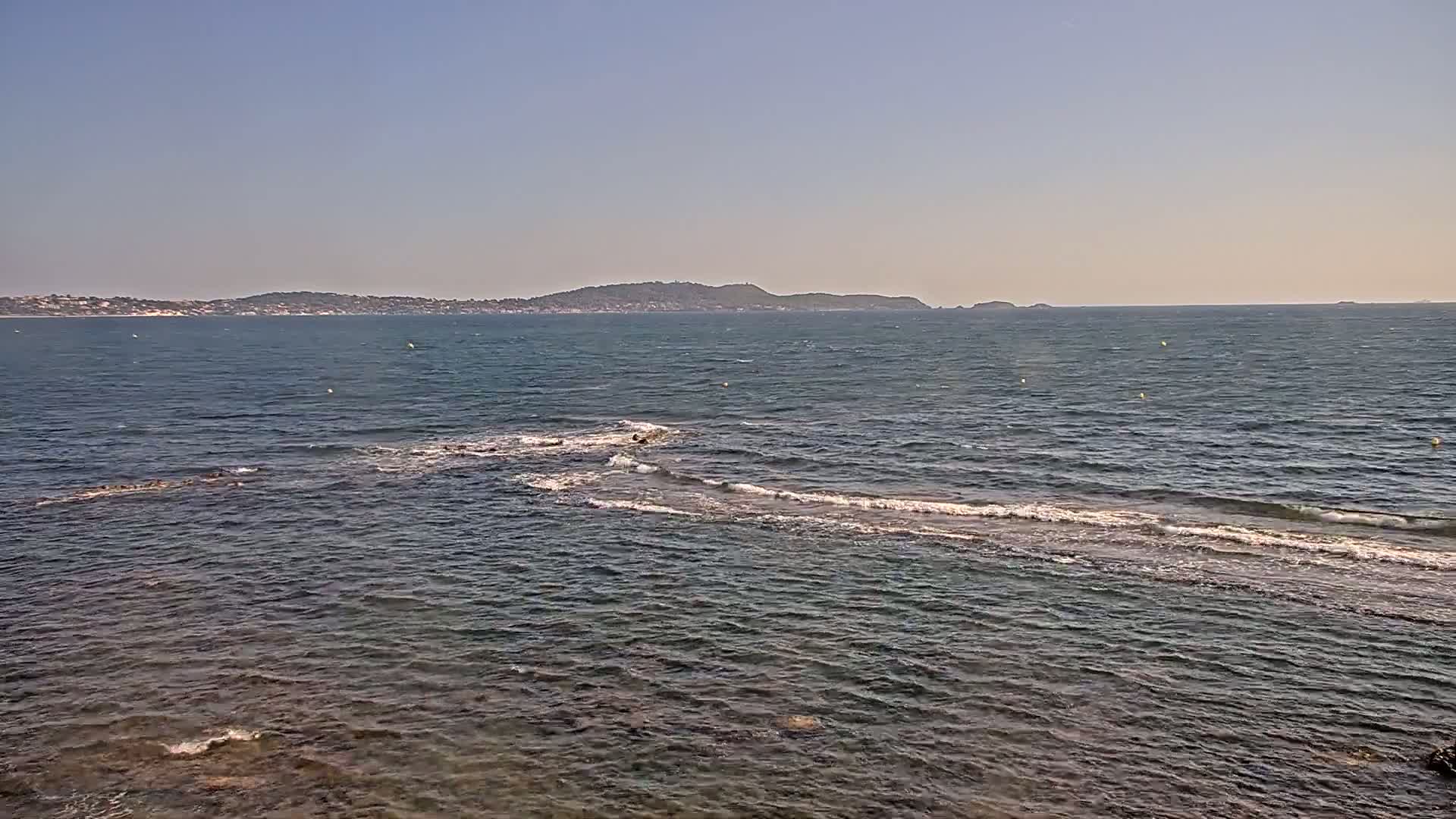 A calm, clear day shows a wavy sea with a distant shoreline and islands under a clear blue sky.