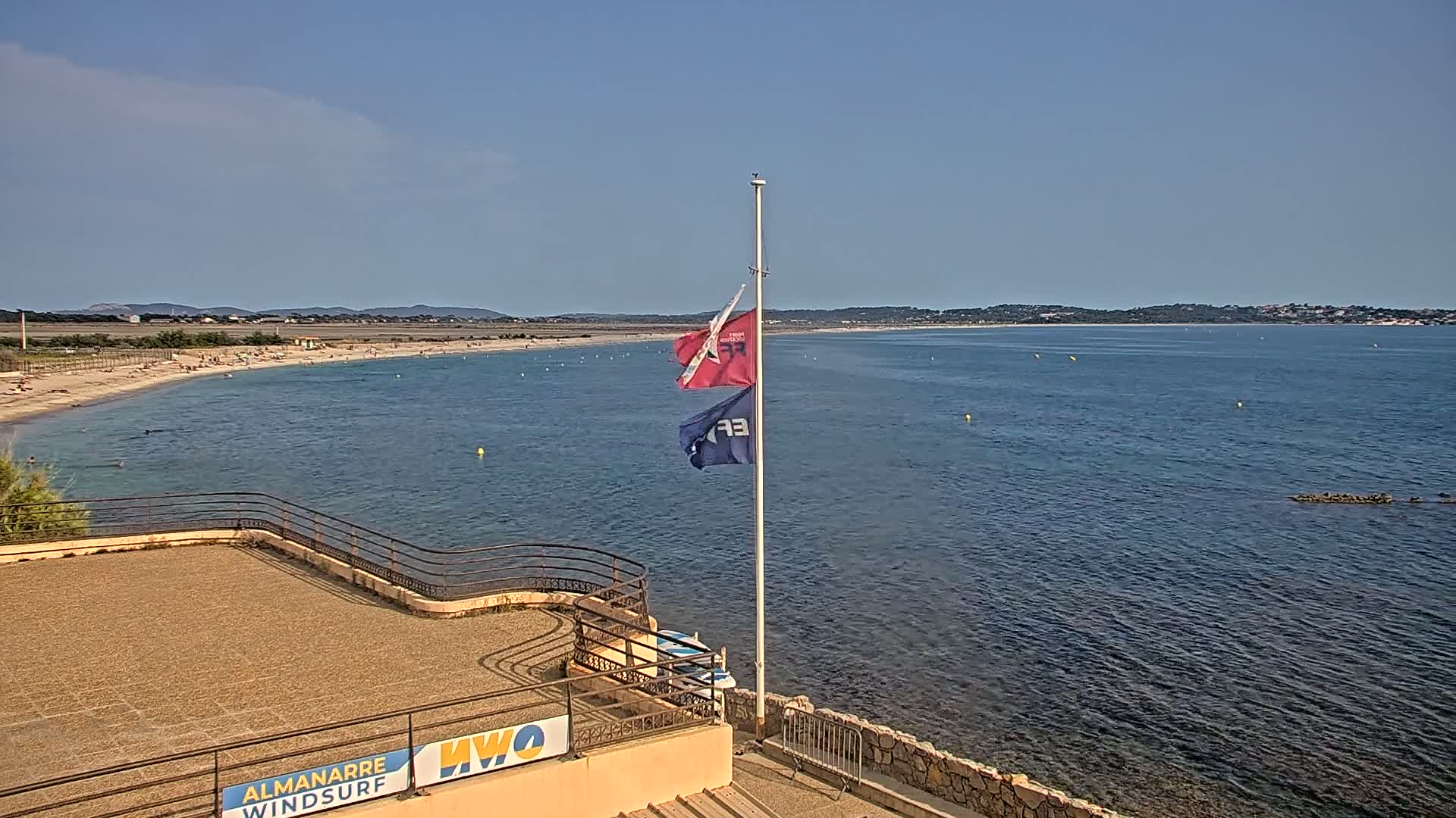 A sandy beach with scattered people stretches along a calm blue sea under a clear sky, with two flags on a pole in the foreground near a stone structure.