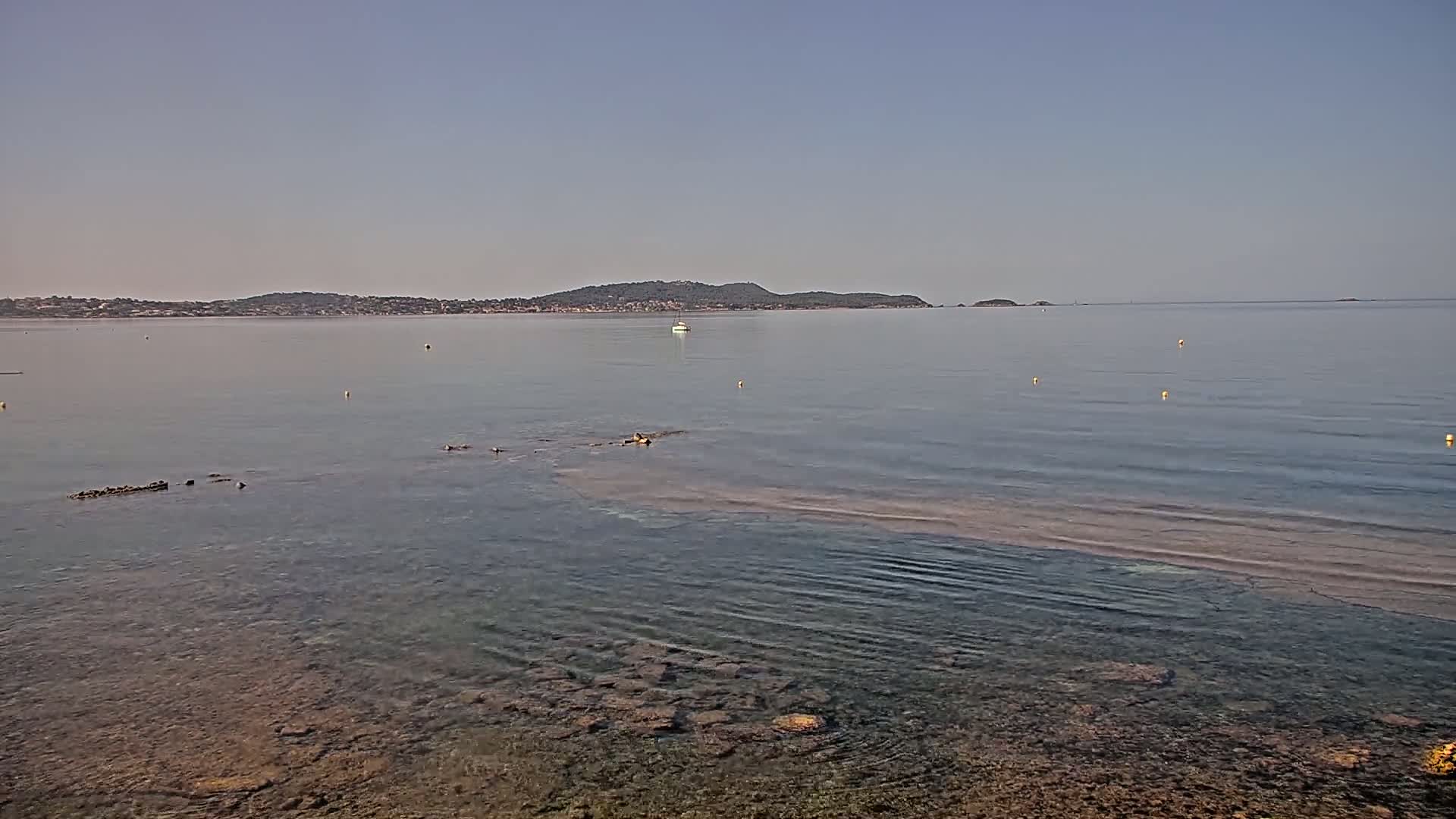 A calm, clear body of water with a rocky shoreline and a distant landmass under a pale sky.