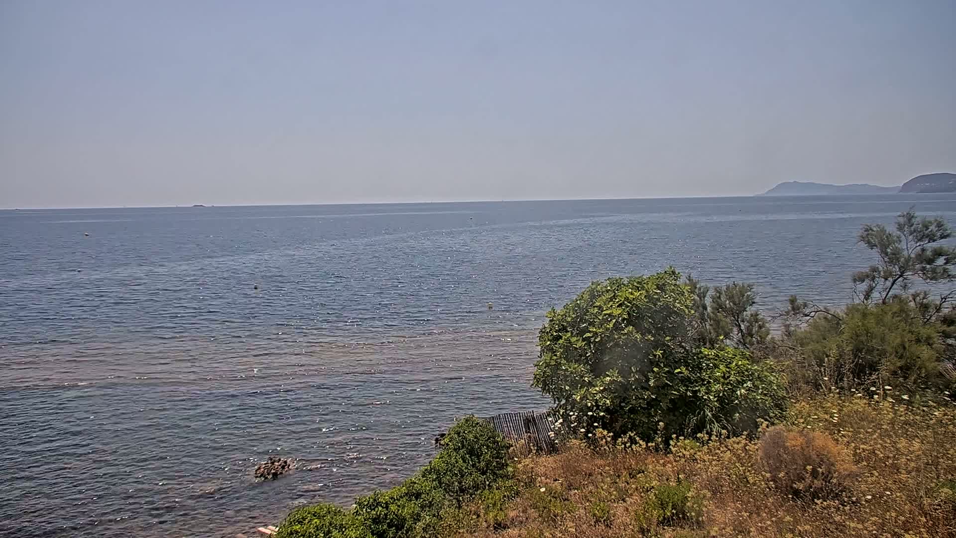 A calm, clear day reveals a view of a tranquil sea extending to distant mountains, with shrubs and vegetation lining the foreground shoreline.