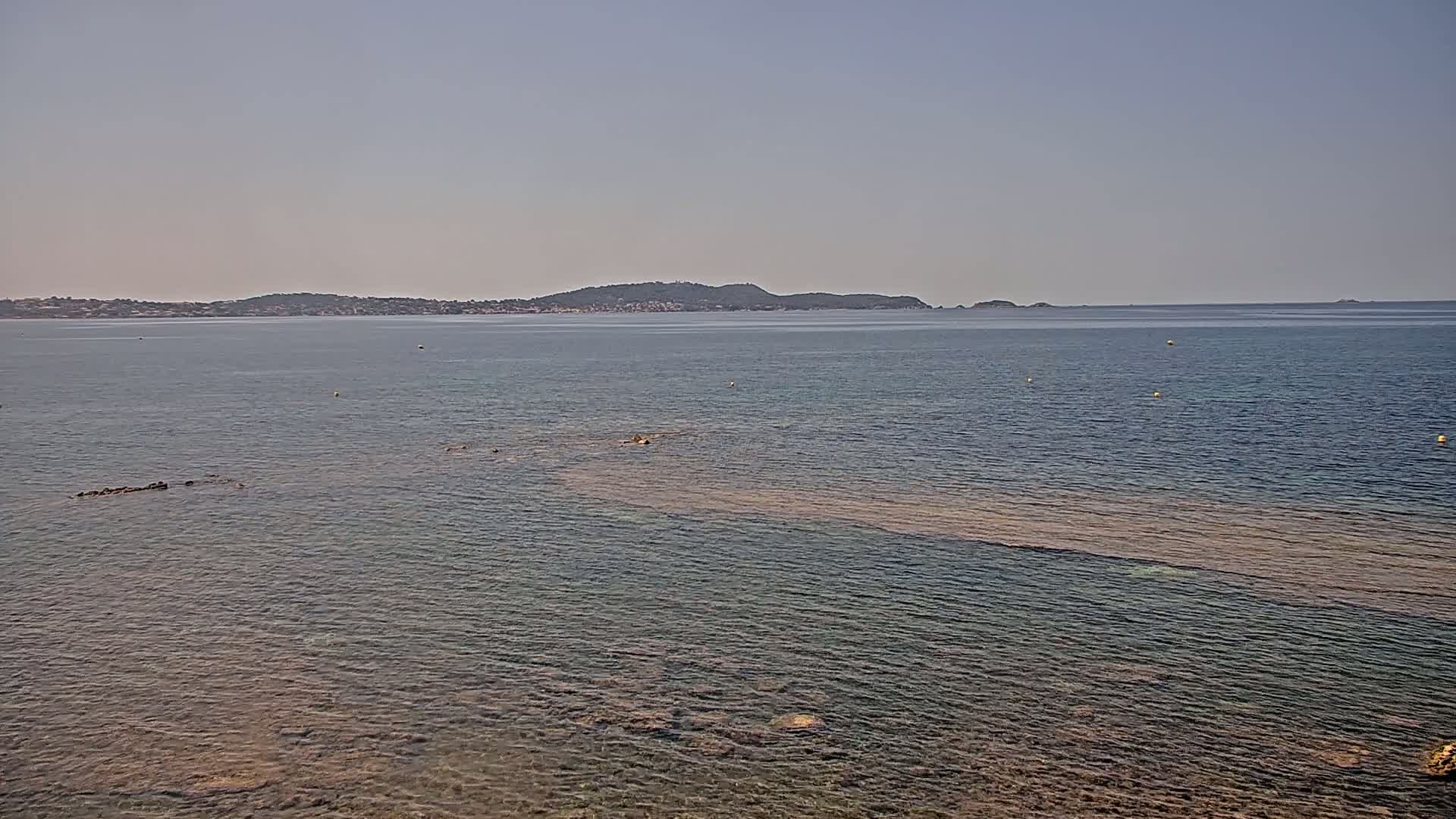 A calm, clear sea with a distant landmass under a hazy sky shows a shallow, rocky area extending from the foreground into the water.