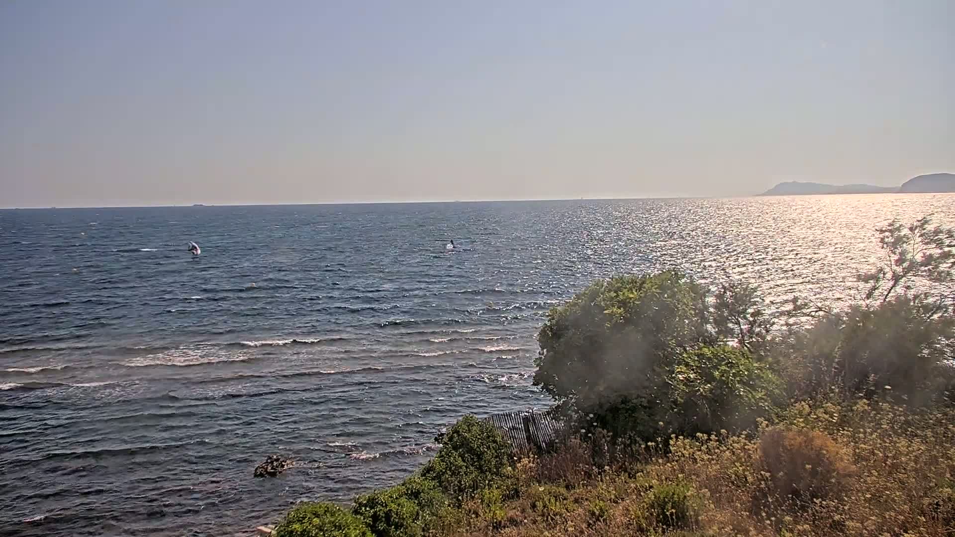 A calm, sunny day reveals a view of a wavy sea with two small sailboats, a rocky shoreline with sparse vegetation, and distant hills under a clear sky.