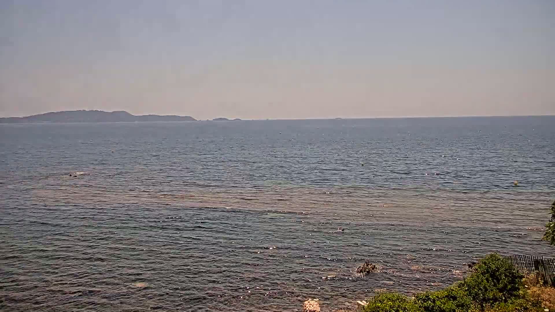 A calm, sunny day shows a view of a body of water with a distant mountain range and a rocky shoreline with green vegetation in the foreground.