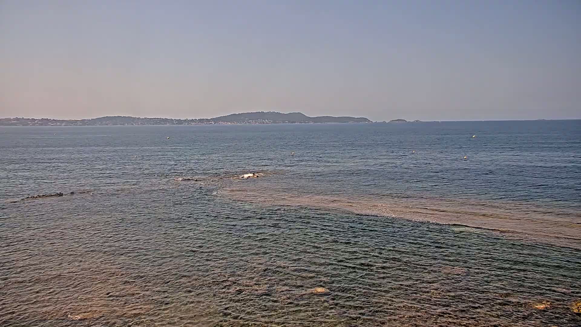 A calm, clear day shows a wide expanse of sea with a distant coastline and a shallow, light-brown area of water closer to the foreground.