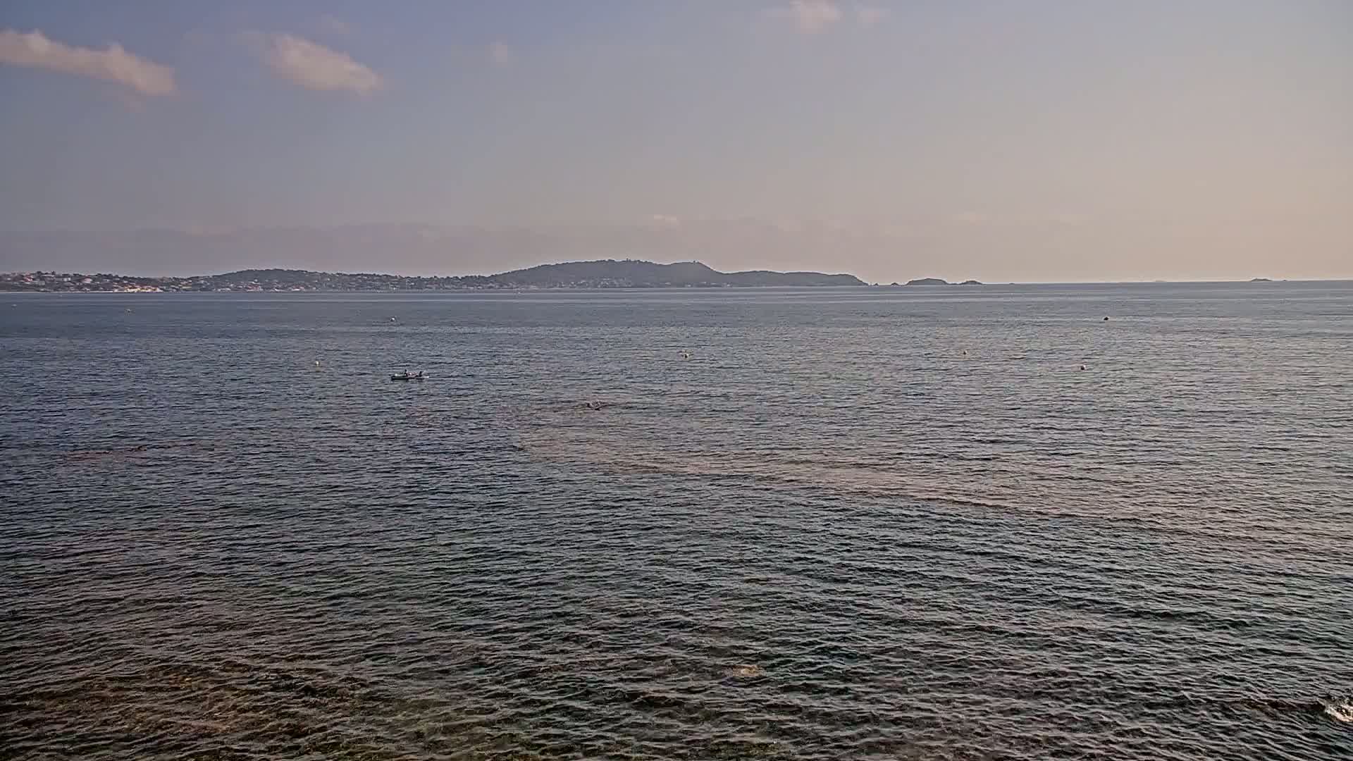 A calm sea under a mostly clear sky shows a distant landmass with a low-lying, tree-covered hill.