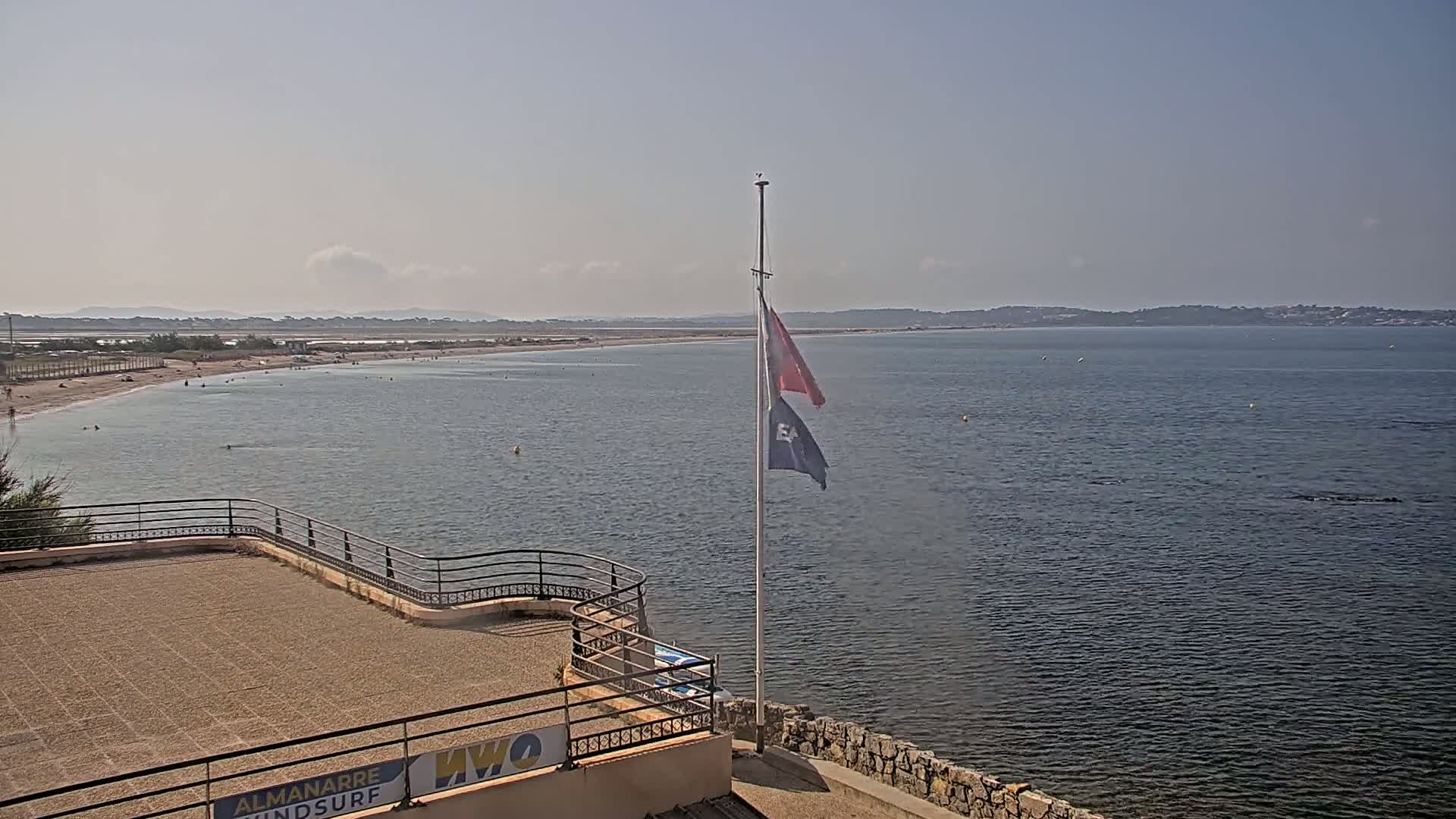 A sandy beach with a few people on it stretches along a calm body of water under a hazy sky, with a flagpole bearing two flags standing on a stone structure overlooking the scene.