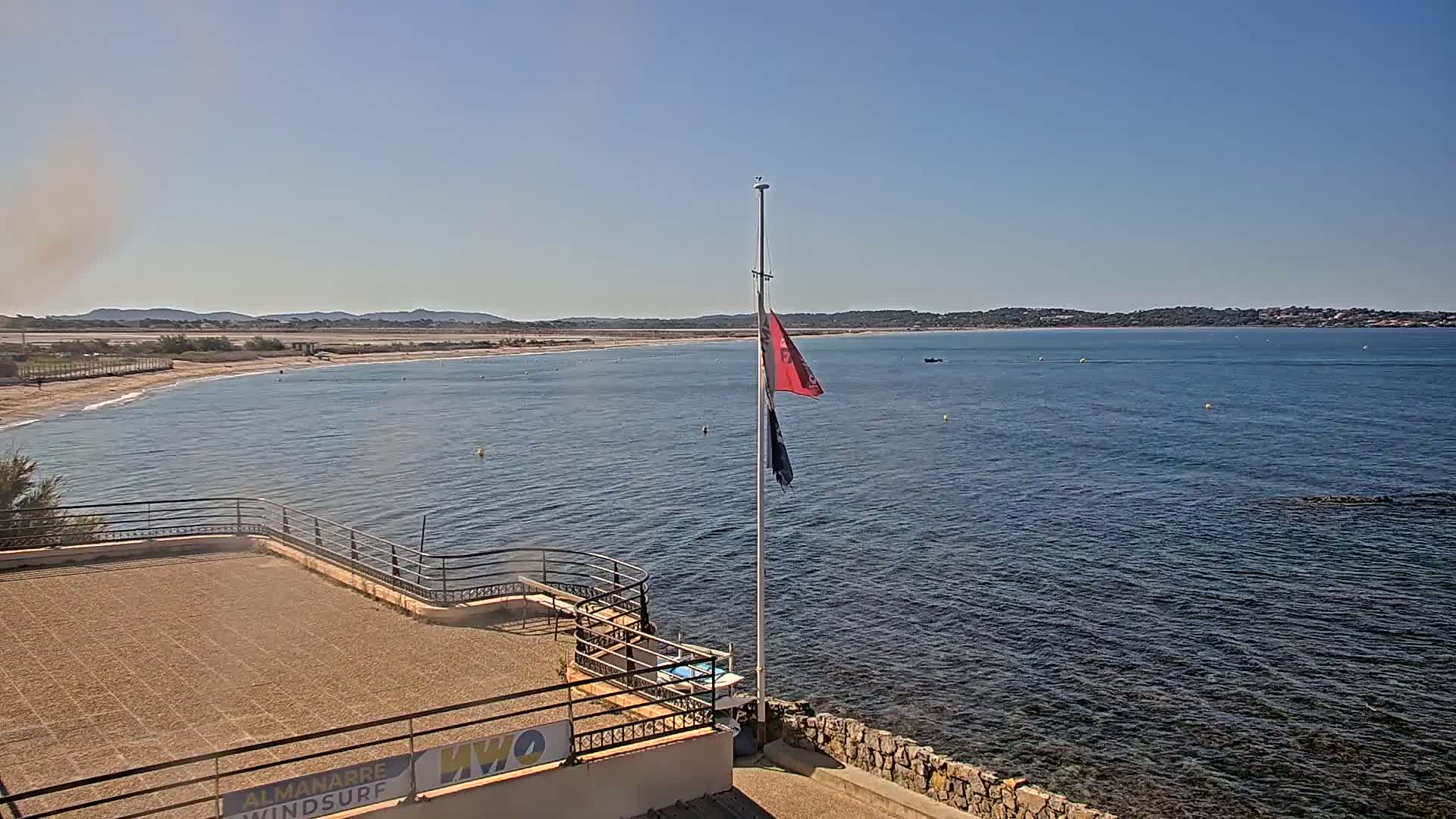 A flagpole with a red and blue flag stands on a stone structure overlooking a calm sea and a sandy beach under a clear, sunny sky.