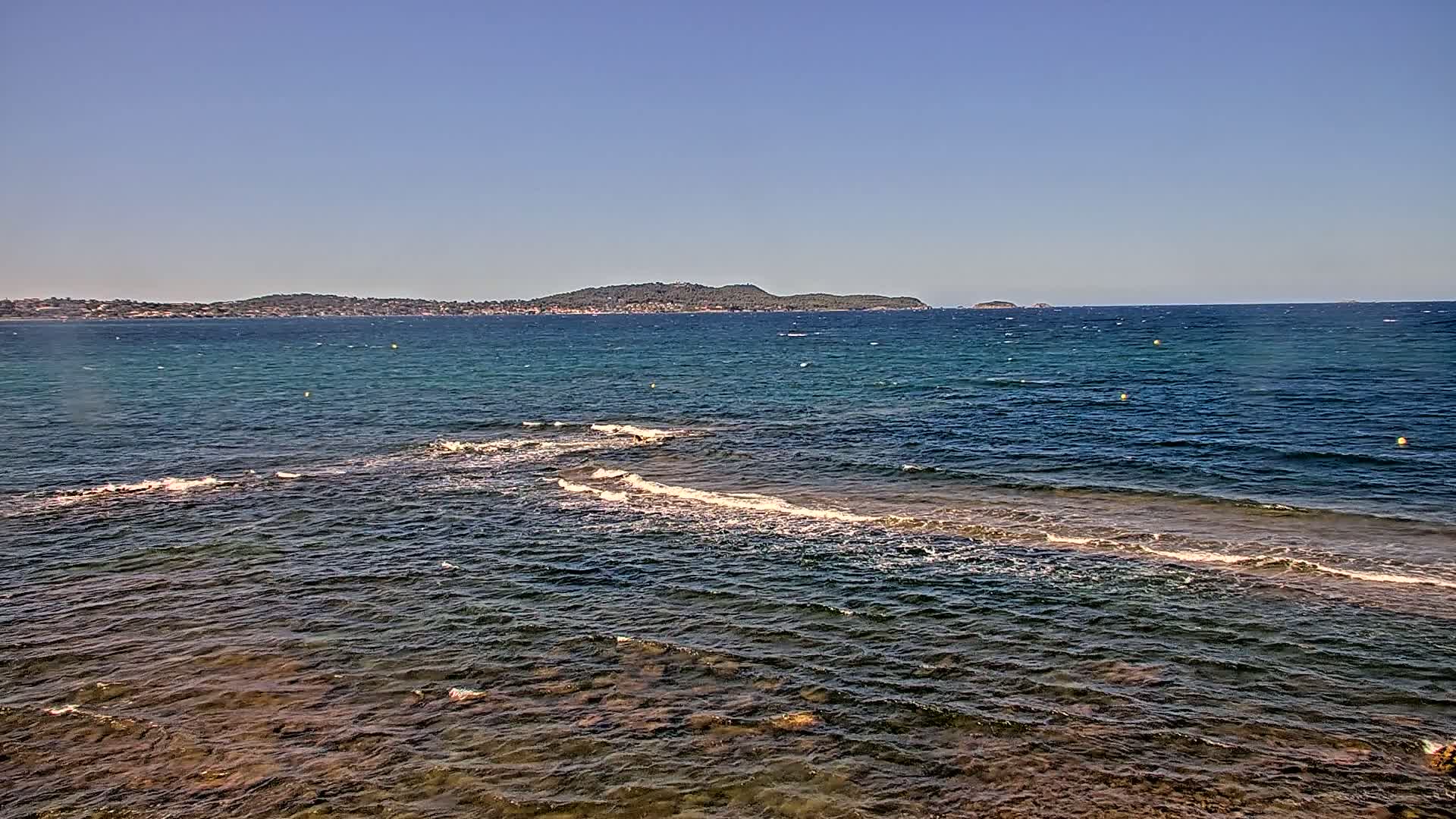 A calm sea with small waves laps against a rocky shore, under a clear blue sky, with a distant island visible on the horizon.