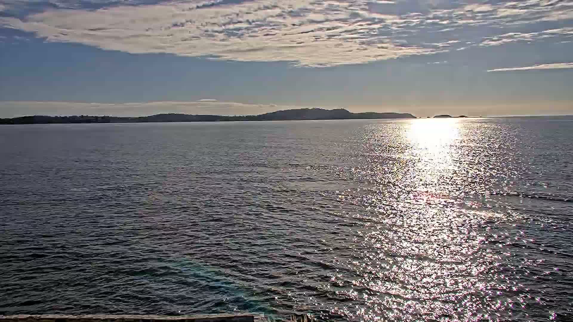 A calm sea with small waves laps against a rocky shore, under a clear blue sky, with a distant island visible on the horizon.