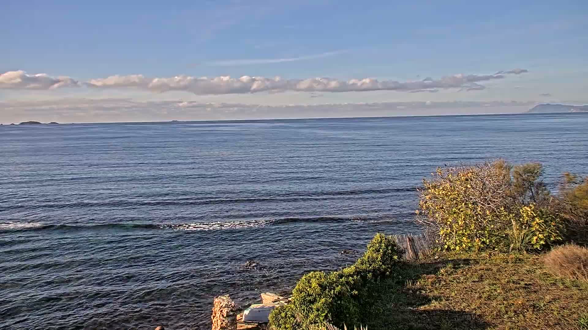 The image captures a sunny, partly cloudy day over a vast blue ocean with gentle waves, viewed from a vegetated hillside, with distant islands and mountains visible on the horizon.