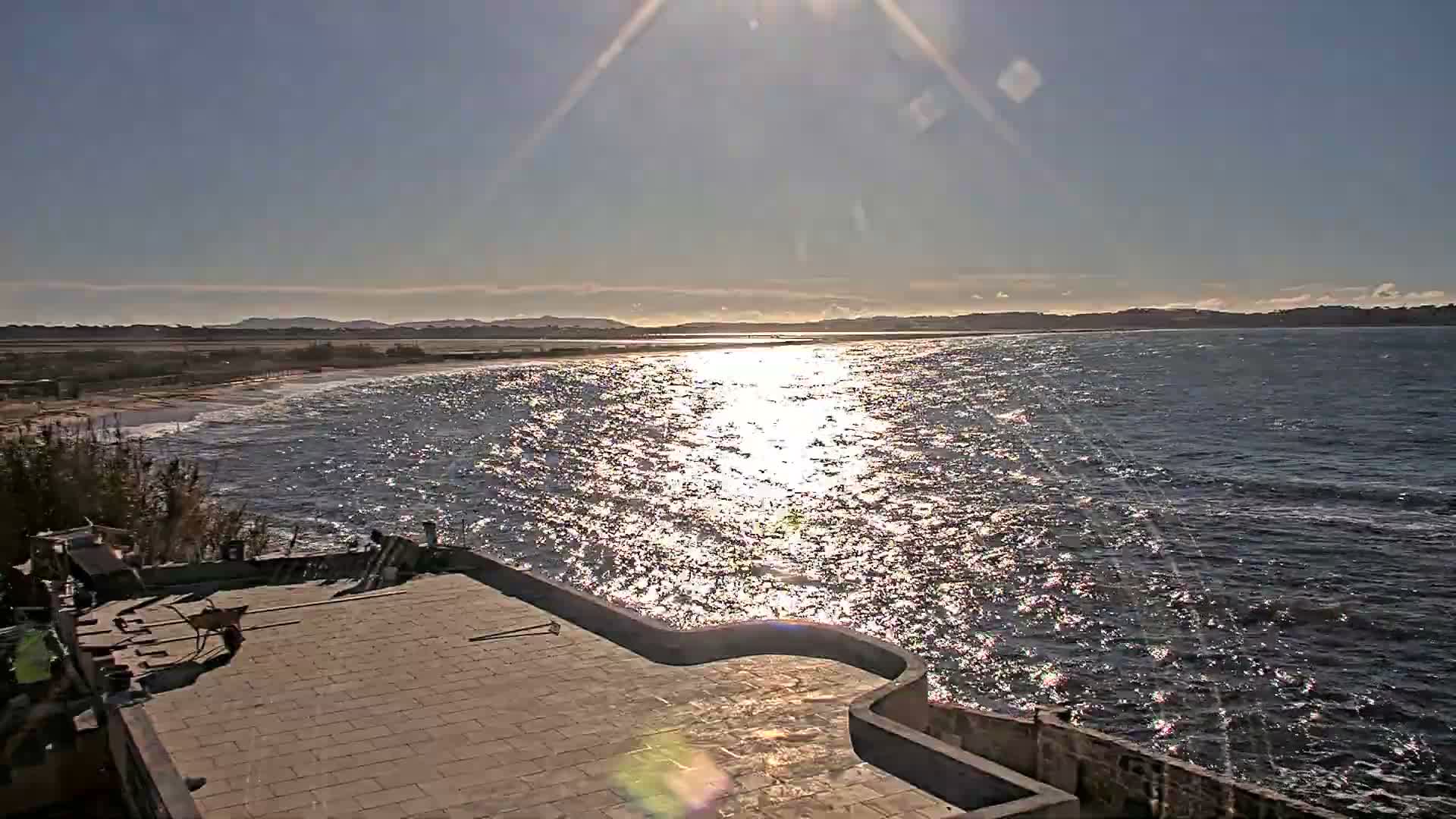 On a bright and sunny day, a wide coastal vista is seen from a paved terrace, revealing a sun-drenched sea with sparkling reflections, a long sandy beach, and distant land.