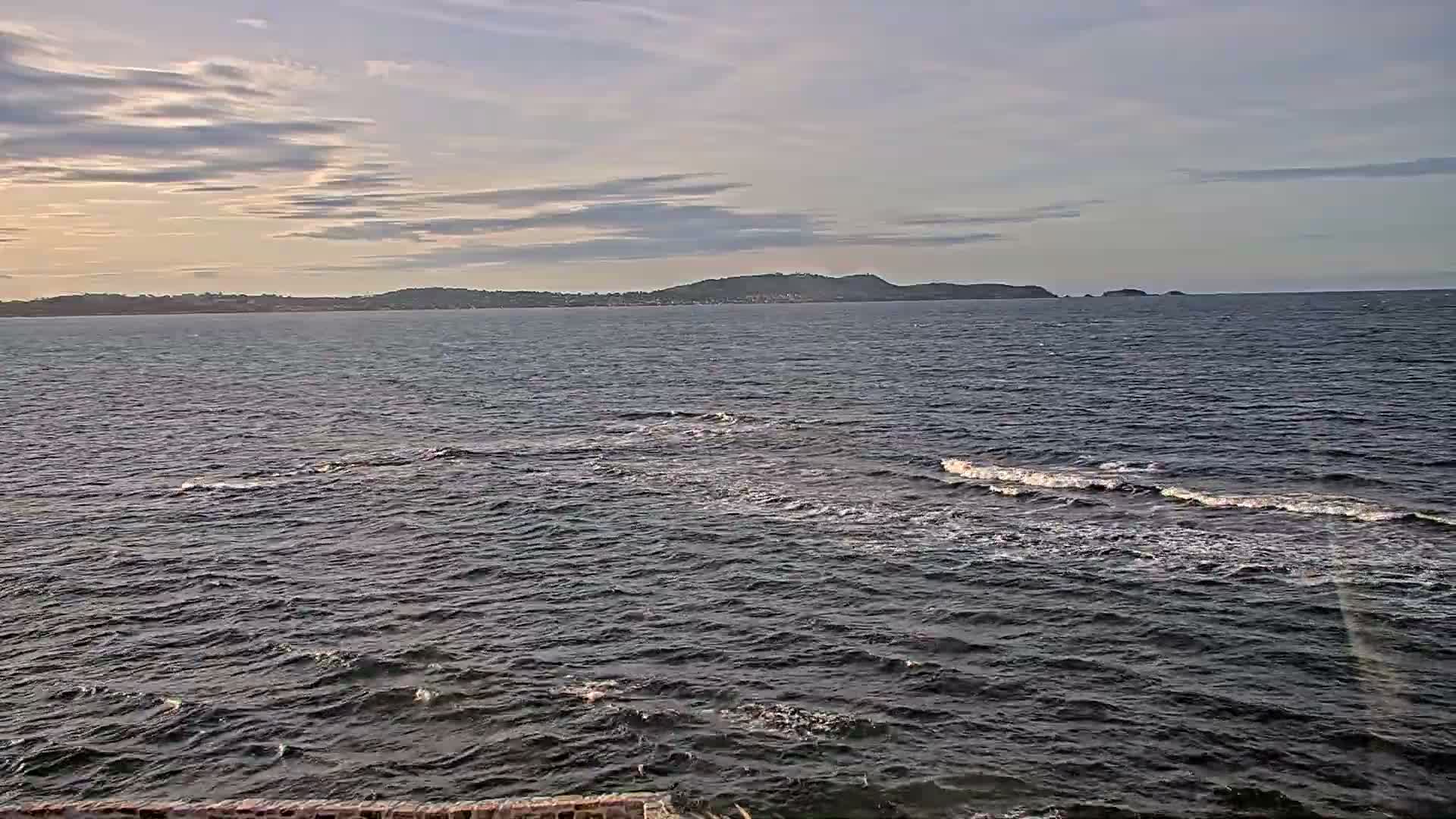 A wide view presents dark, choppy ocean water with visible waves breaking into white foam, extending to a distant hilly coastline under a partly cloudy sky with a warm glow on the left horizon.