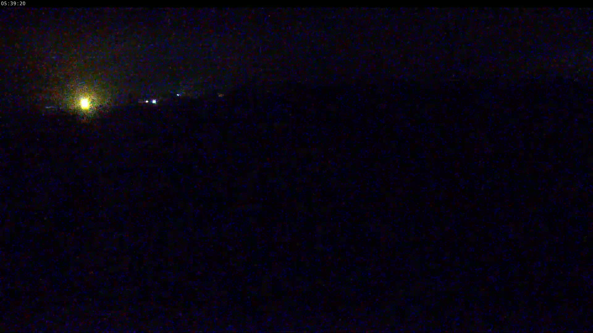 A dark, nighttime scene shows a bright light in the distance, possibly from a vehicle's headlights, and other faint lights, suggesting an area of low-level illumination.