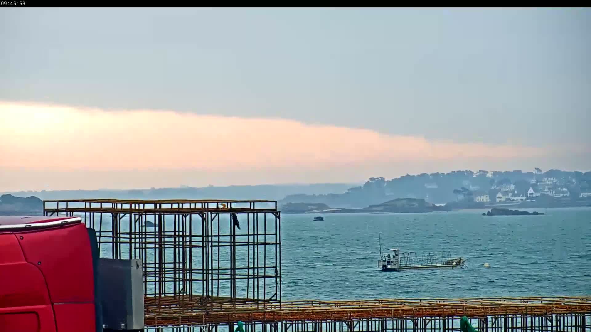 A red truck is partially visible in the foreground next to a metal framework overlooking a calm body of water under a pale pink and gray sky, with a small boat and distant coastline in the background.