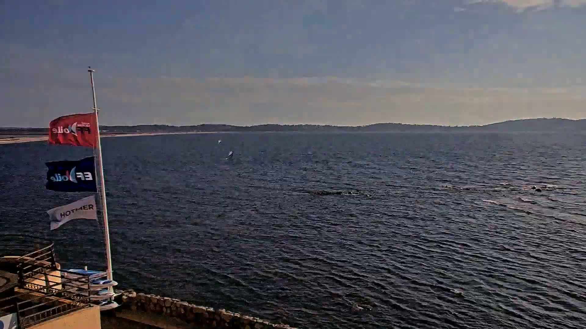 A calm body of water with a distant shoreline is viewed from a vantage point near a flagpole displaying several flags and some small watercraft.