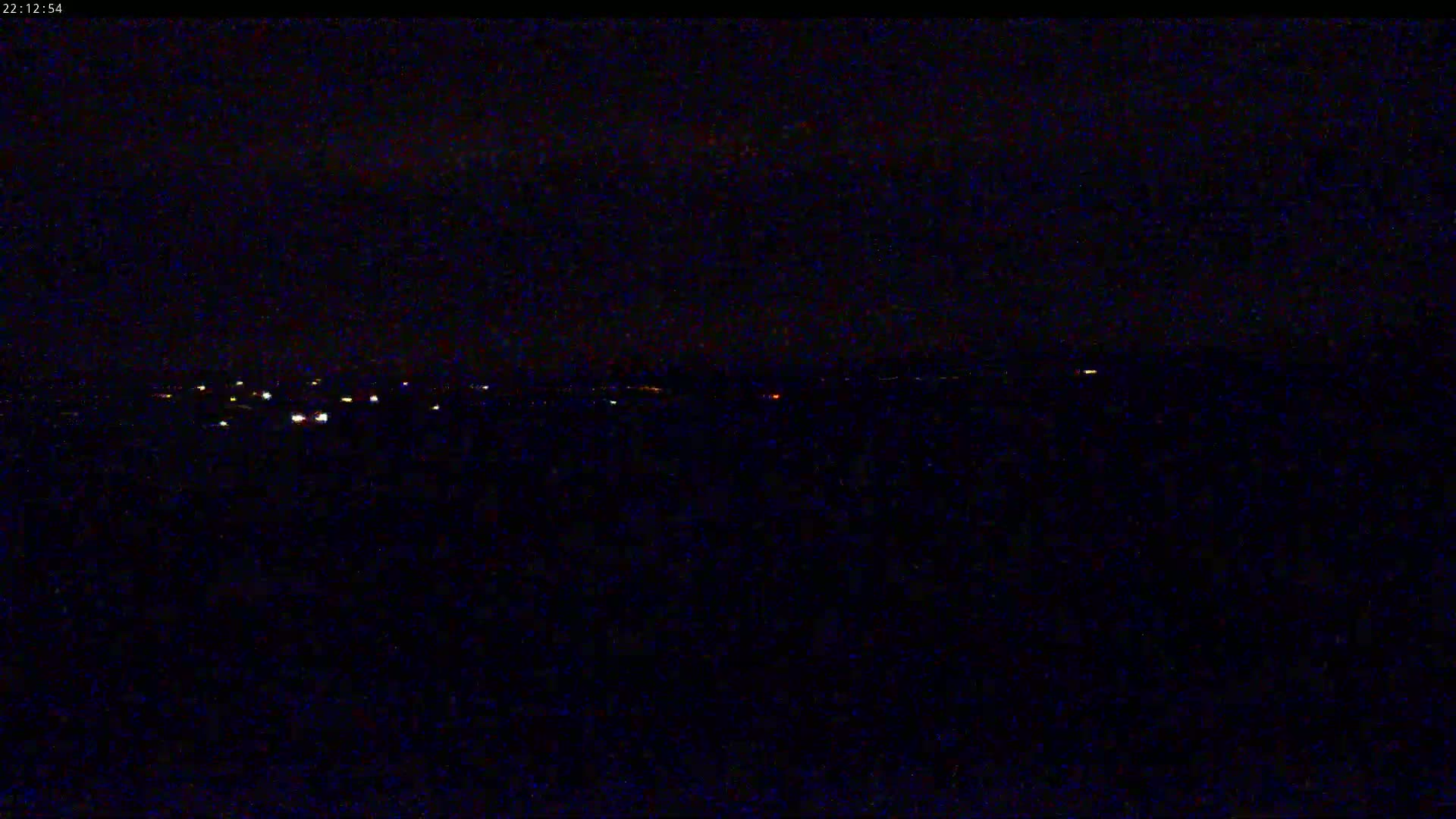 A dark, nighttime landscape shows a distant line of faint lights on the horizon under a mostly clear sky.