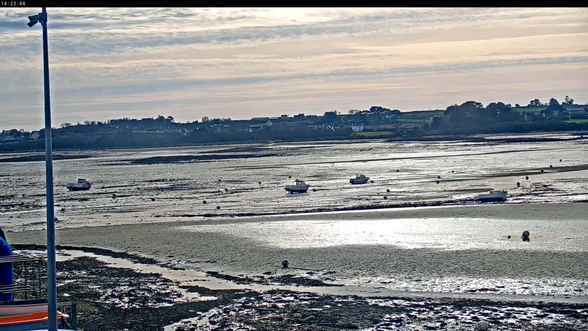 A mostly sunny day shows a wide expanse of mudflats at low tide with several small boats resting on the exposed seabed, a distant village visible on the horizon.