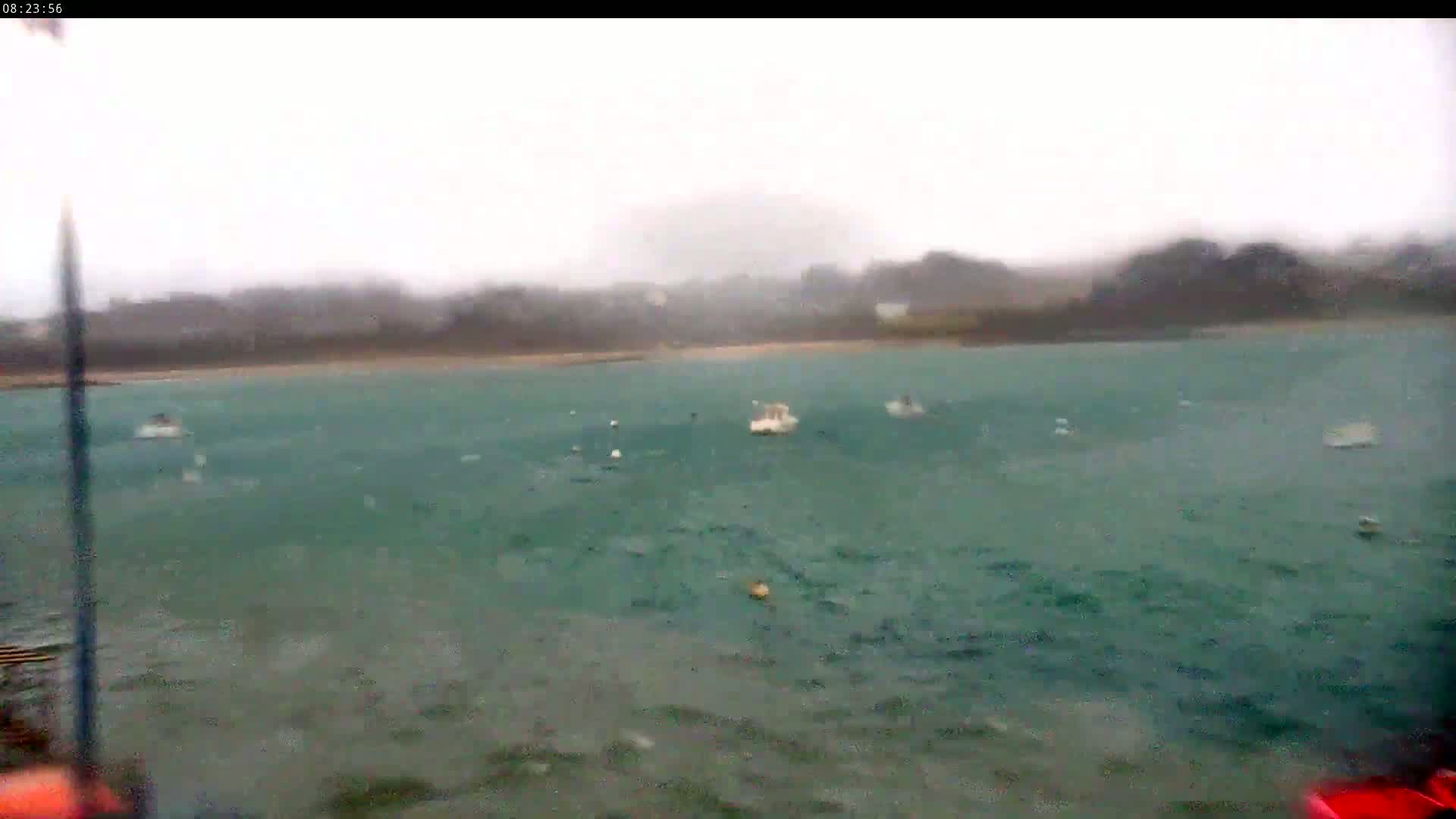 A choppy, teal-colored body of water with several small boats visible under an overcast sky during a rainstorm.