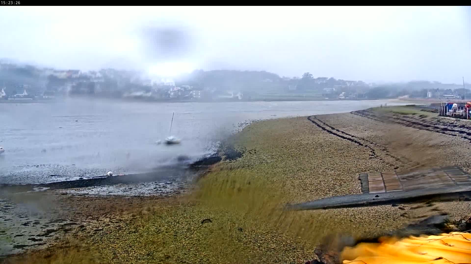 A misty, overcast day reveals a shoreline with a gravelly beach, a calm body of water, and a small boat in the distance.