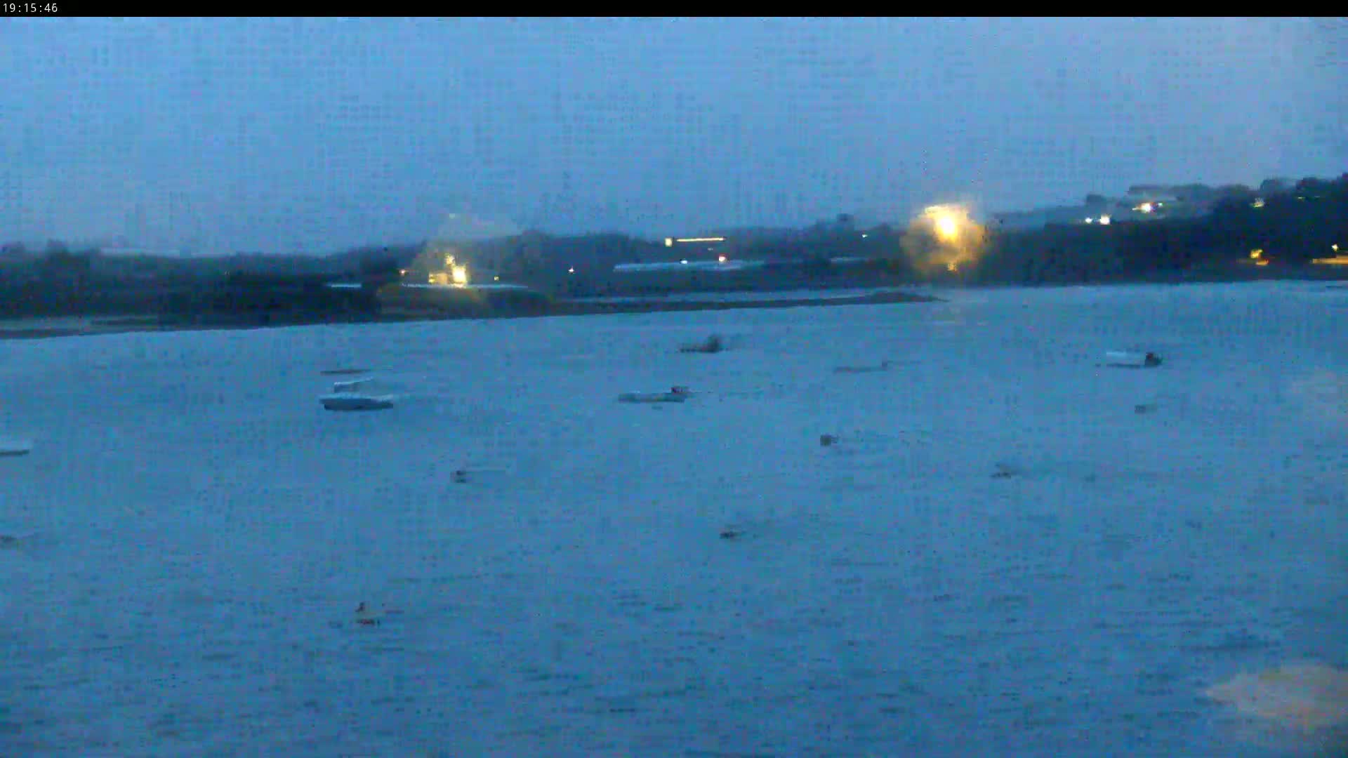 Several small boats are scattered across a dark, calm body of water at dusk, with a shoreline and some lights visible in the distance.